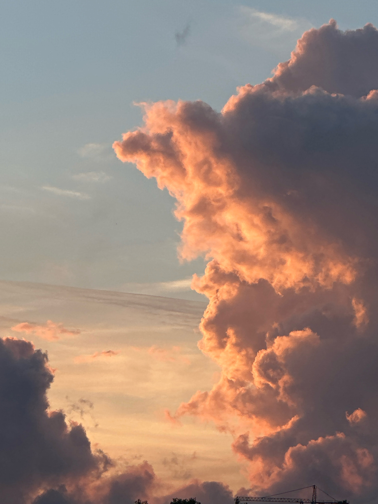 The image is a close-up, eye-level shot of the sky filled with dramatic clouds during sunset. The clouds are a mix of deep purples, grays, and vibrant oranges and pinks, illuminated by the setting sun. The sky transitions from a pale blue at the top to a warmer, more muted orange hue lower down. In the bottom right corner, the latticed structure of a construction crane is visible against the sky, suggesting an urban or industrial setting. The scene is devoid of any people. The image captures the atmospheric beauty of a sunset over Tallinn, Estonia, highlighting the interplay of light and shadow on the clouds and hinting at the city's skyline or construction projects.
