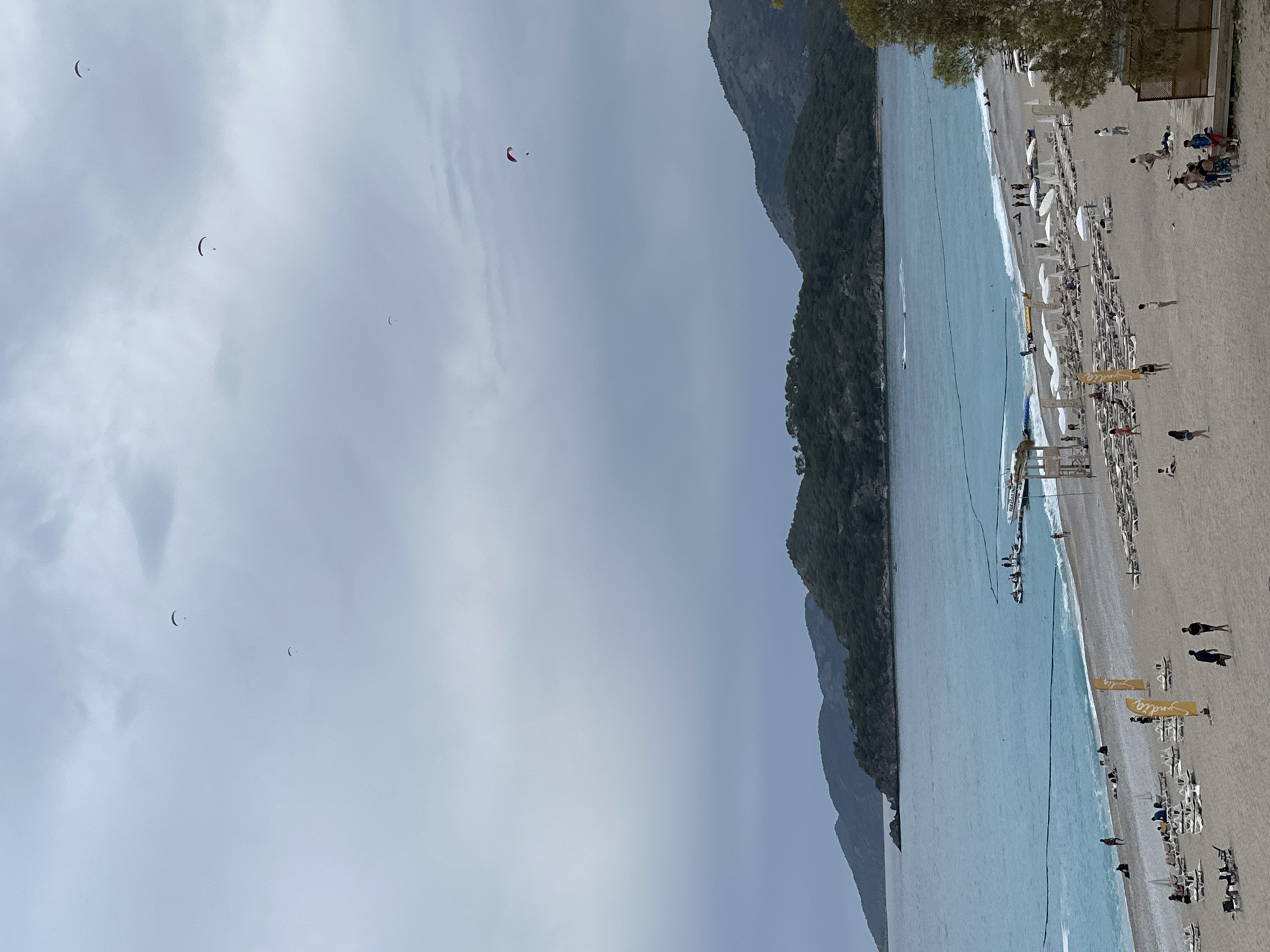 The image shows a beach in Fethiye, Turkey, likely Oludeniz. The scene includes a sandy beach with sunbeds and umbrellas, the Mediterranean Sea, mountains, and a partly cloudy sky. People are seen walking and relaxing on the beach. There are several paragliders in the air.