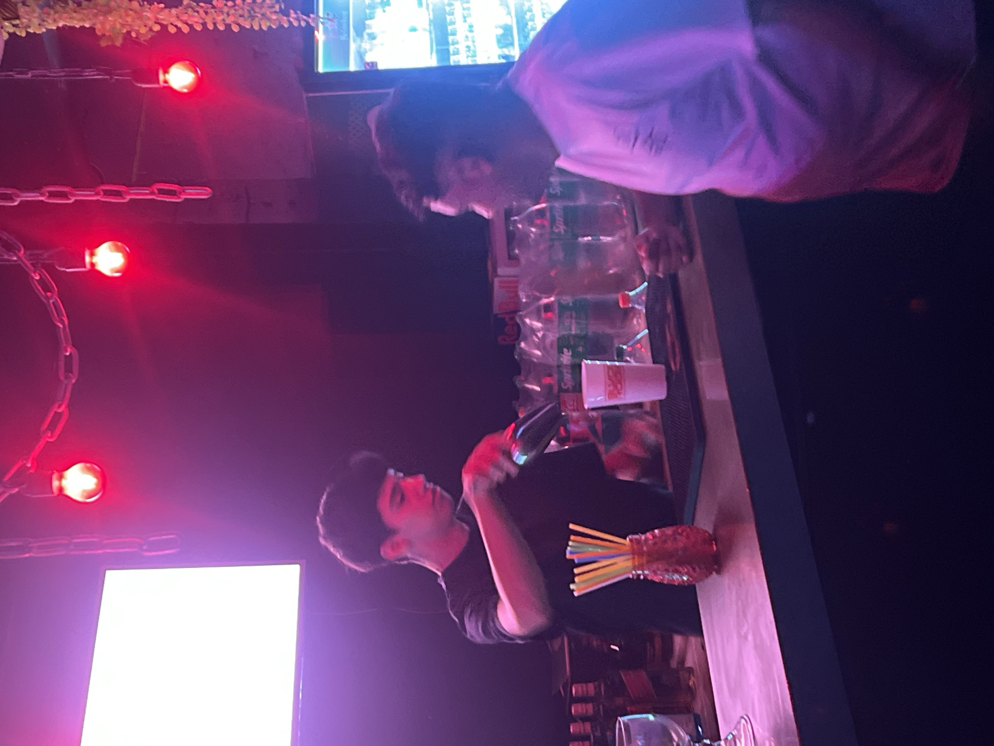 Two people stand behind a bar. One is holding a bottle in their hand. There are drinks stacked behind the bar. The lighting is red. There are chain lights in the background. There is a sign on the left. The city is not mentioned. The country is not mentioned.