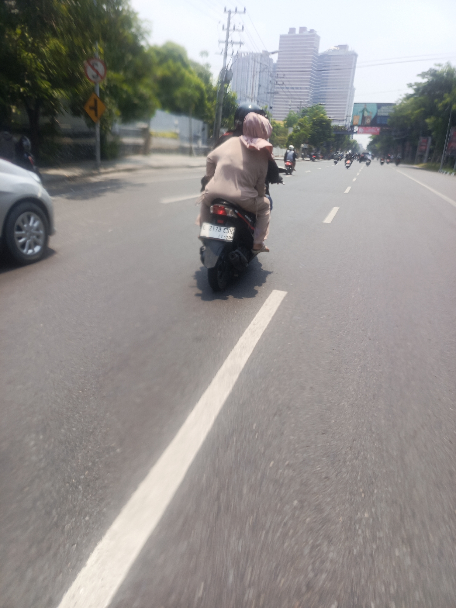 The image depicts a street scene in Surabaya, Indonesia, likely on a sunny day. In the foreground, a person is riding on the back of a motorcycle, wearing a light brown hijab and clothing. The motorcycle has a license plate that appears to read "L 2178 CBH". To the left, the front of a silver car is visible. The road has white dashed and solid lines. In the background, several other motorcycles are also on the road, moving in the same direction. Further in the distance, tall buildings, possibly skyscrapers, rise above the trees, and a billboard is visible. Traffic lights and utility poles are also present, indicating an urban environment. A traffic sign with a red circle and a horizontal line, likely indicating "no stopping" or "no parking," is visible on the left side of the road, along with a diamond-shaped yellow sign with directional arrows. The overall impression is of moderate to heavy traffic on a city street.