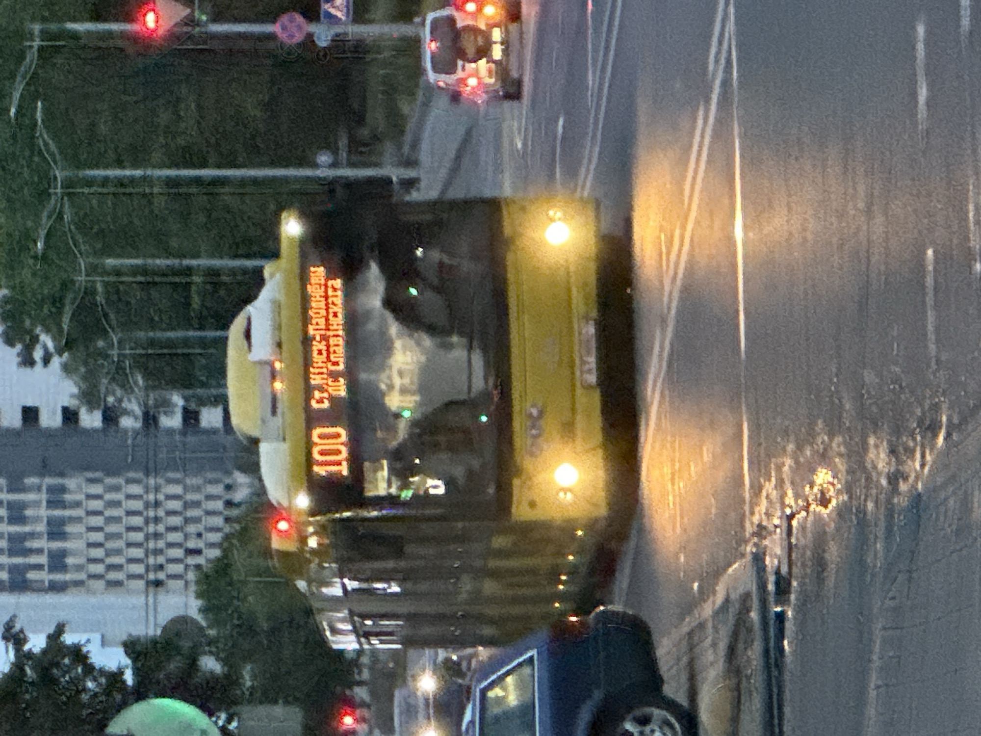 This image captures a street scene in Minsk, Belarus, likely during dusk or early night, given the illumination of vehicles and streetlights against the ambient light. The central subject is a yellow articulated trolleybus, displaying route number "100" and destinations "Ст.Минск-Паўднёвы" (Minsk-South Station) and "ДС Славінскага" (Slavinskogo Bus Station), confirming the vehicle's operation within Minsk. The bus is oriented towards the viewer, slightly angled to the right, with its headlights and interior lights on, and its destination sign brightly lit.
To the immediate left of the bus, a dark blue car, possibly an older sedan, is partially visible from its rear left side, with its wheel and fender in the foreground. Further down the street on the right, a white SUV, possibly a UAZ Patriot, with a spare tire mounted on its rear door, is visible with its taillights illuminated.
The road surface appears wet, reflecting the bright lights from the vehicles, suggesting recent rainfall. White road markings are visible on the asphalt. Red traffic lights are prominent on both the left and right sides of the frame, indicating a stop or intersection ahead. A green traffic light is partially visible on the far left. In the background, beyond the bus and trees, a modern building with a distinctive checkered facade of light and dark squares is visible. Streetlights with multi-arm designs are also present on the right side of the road. No distinct individuals are clearly visible or performing specific actions, though faint reflections of occupants can be seen in the bus windshield.