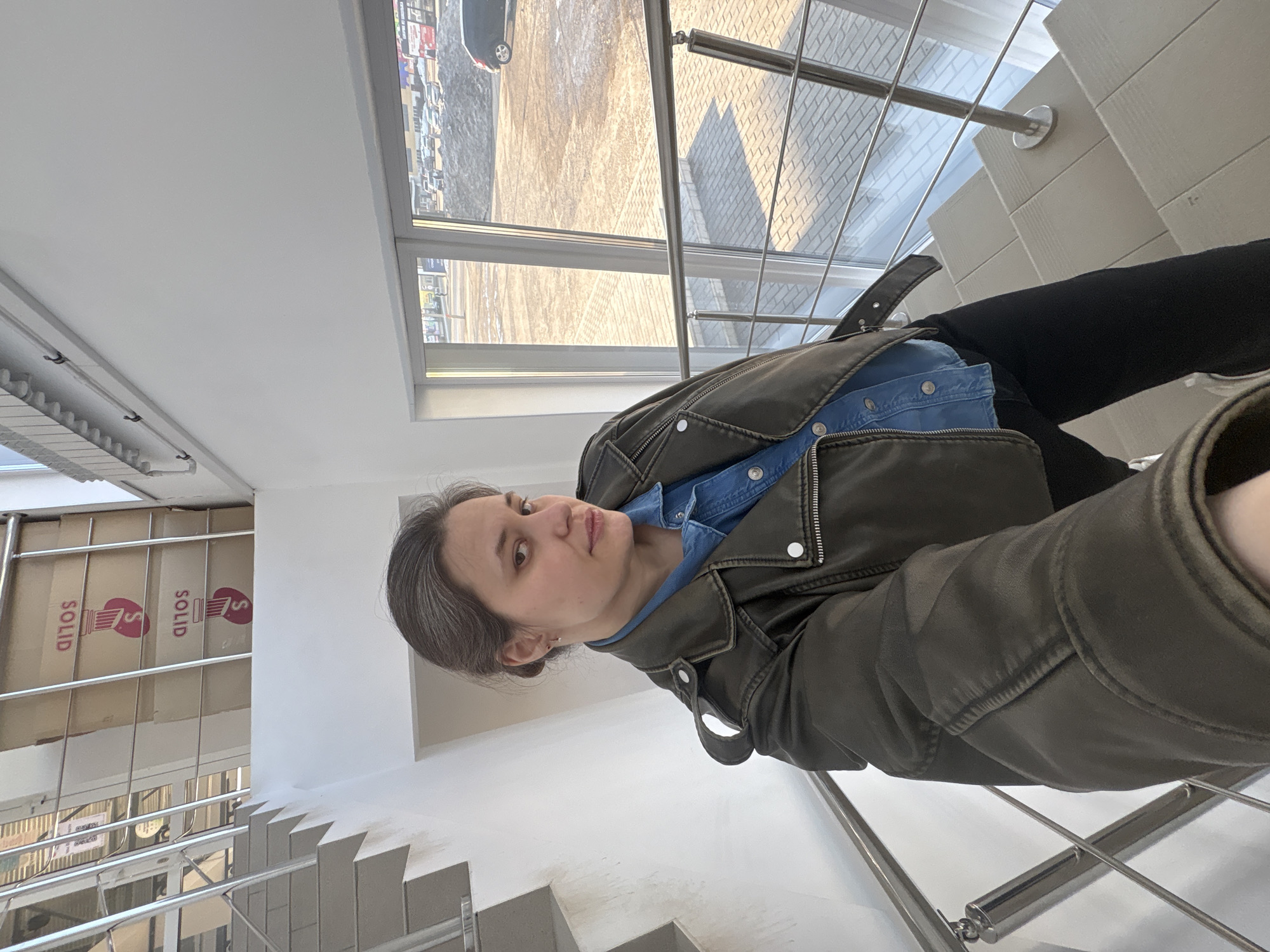 A woman stands on stairs with a silver railing inside a building. She is wearing a blue denim shirt and a khaki jacket. Boxes with "SOLID" printed on them are visible on the left, stacked near the staircase. Outside the building, a parking lot and a multi-story building are visible through a window. Based on visual clues and the request for location in Chaikovsky, Russia, it is possible that this building is associated with the Solid shopping center.