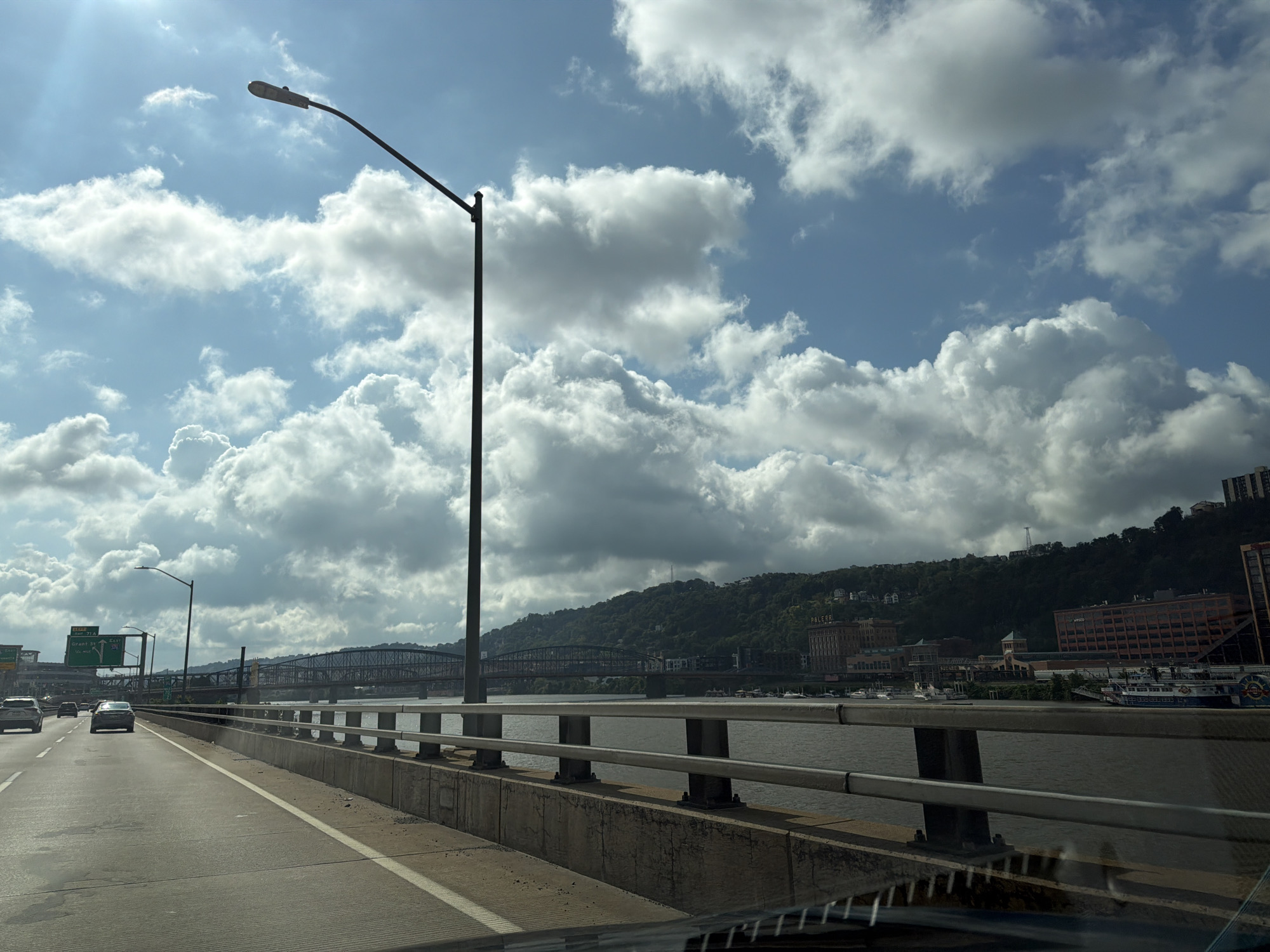 An eye-level, wide-angle shot captures a view from a highway bridge in Pittsburgh, Pennsylvania. The sky is dominated by a mix of bright sunlight and dramatic, white cumulus clouds, suggesting a fair-weather day. A tall streetlamp on the left side of the road is silhouetted against the sky. The highway stretches forward, with several cars visible, indicating moderate traffic. To the right, a river flows, bordered by a concrete barrier with metal railings. In the distance, a city skyline emerges, featuring bridges crossing the river and buildings nestled against a lush, tree-covered hillside. Some of these buildings appear to be residential, while others are larger commercial structures. A large, multi-deck boat is docked on the riverfront. The overall impression is a bustling urban landscape under a dynamic sky.