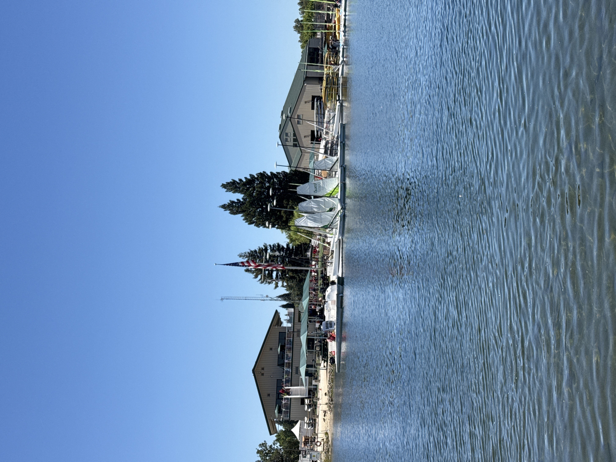This is a bright, sunny, eye-level, medium shot of a lake with a building and sailboats on the shore. The lake takes up the bottom half of the image, and the water is clear enough to see the sandy bottom in some areas. The shore features a long, light-colored building with a green roof, several sailboats moored nearby, and some kayaks stacked on the sand. An American flag flies from a flagpole in front of the building. Several trees are visible behind the building and to the right. The sky is clear and blue. The image appears to be taken at Hagan Community Park in Rancho Cordova, California, a popular spot for watersports and recreation.