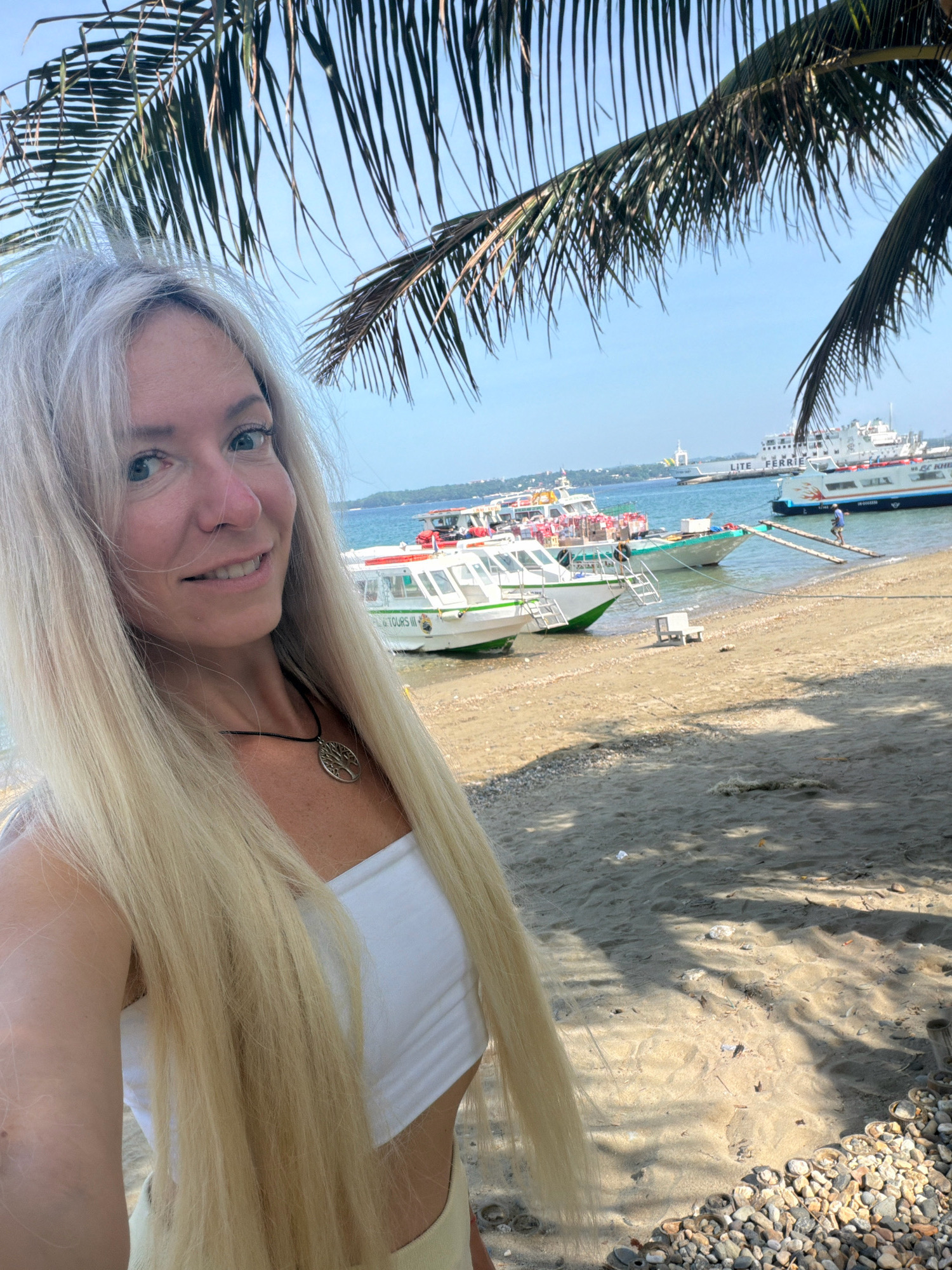 A medium shot captures a blonde woman with blue eyes and fair skin standing on a beach in Malay, Philippines. She is wearing a white tube top and a necklace with a tree pendant. Her long blonde hair is flowing. Behind her, the ocean is visible with several boats docked. One boat has "TOURS III" written on its side. In the distance, larger ferries are anchored. Palm fronds hang from the top of the frame, casting shadows on the sand. The beach is a mix of sand and small pebbles.