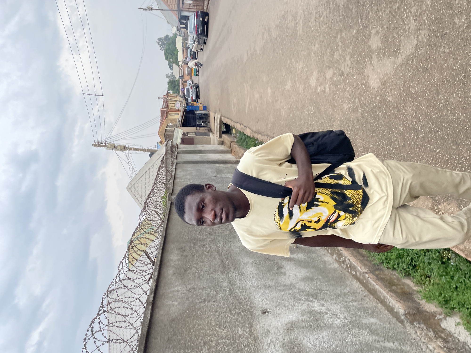 A young man stands on a street in Jos, Nigeria. The street has parked cars and several buildings in the background. A barbed wire fence is to the left of the man. The sky is overcast. The man is facing the camera and is wearing light-colored pants, and a matching shirt with a print of a yellow face. He is wearing a black bag slung over his left shoulder.