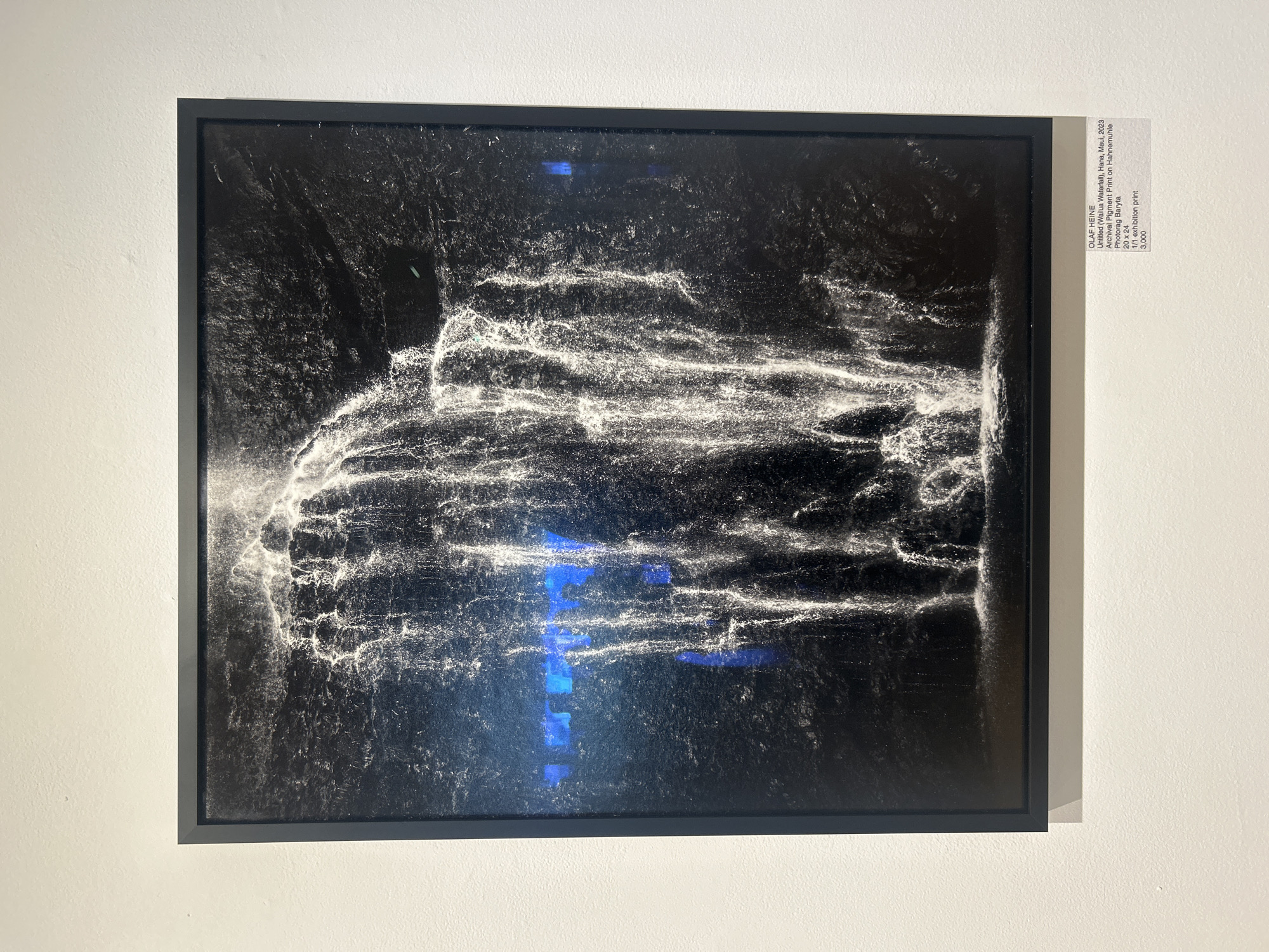 The image shows a framed piece of art hanging on a white wall. The artwork is a black and white photograph with an abstract, textured appearance. There are areas of bright white swirling patterns against a dark background. There are also blue, rectangular reflections on the surface of the photograph, presumably from lights in the room. A small label is affixed to the wall next to the artwork. The label identifies the piece as "Untitled (Wailua Waterfall), Hana, Maul, 2023" by Olaf Heine. It further states that it is an archival pigment print on Hahnemuhle Photorag Baryta, measures 20 x 24, and is a 1/1 exhibition print, priced at 3,000. The artwork's subject matter is Wailua Waterfall, located in Hana, Maui, which is not in West Hollywood.