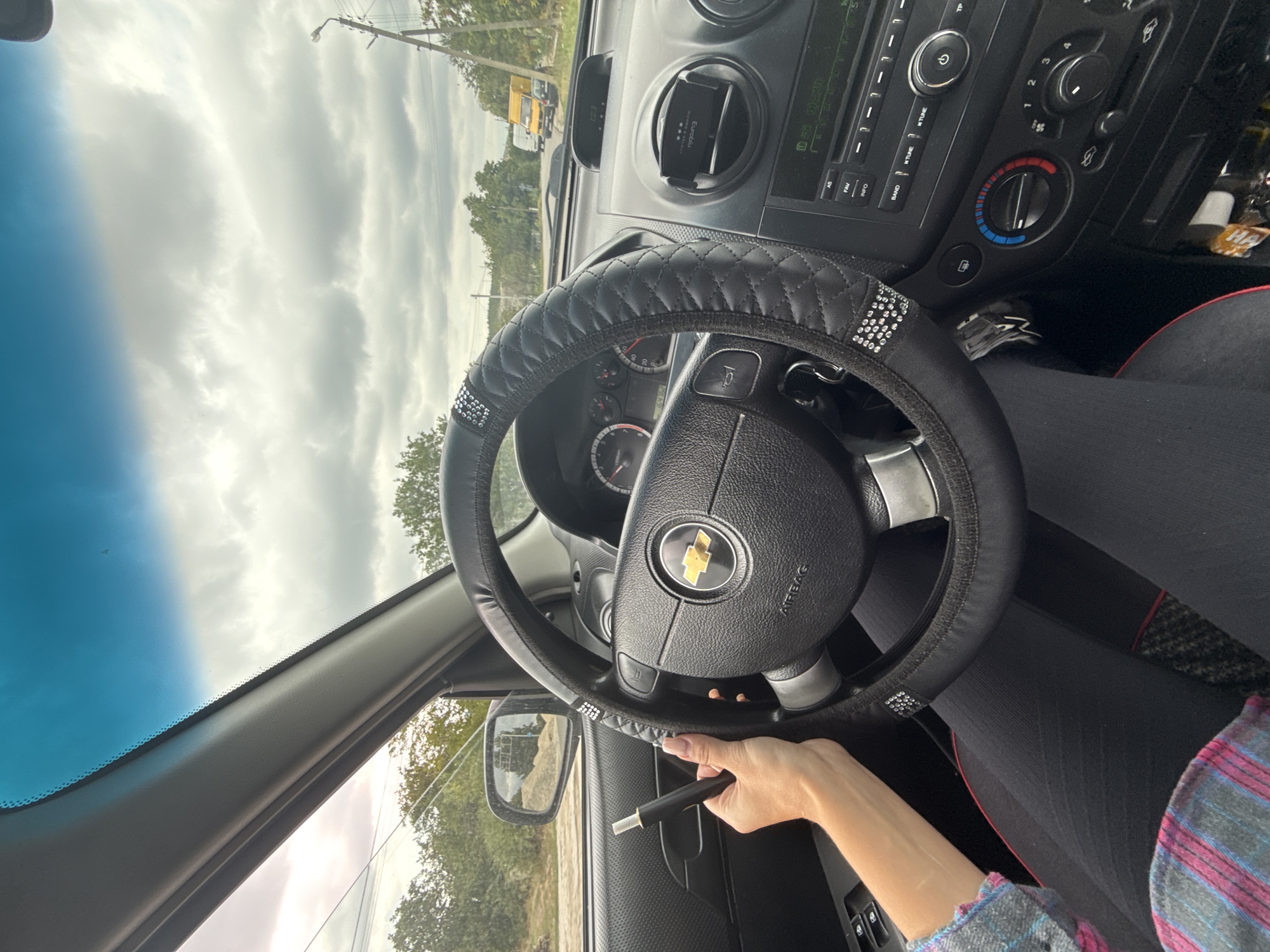 The image shows the interior of a car, taken from the driver's perspective. A person's left hand is visible on the left side of the steering wheel, holding a black electronic cigarette. The steering wheel has a black quilted cover with rhinestones. The dashboard and center console are visible, including the instrument cluster, air vents, and a car stereo. The car is driving on a paved road, with trees and power lines visible outside the windows. The sky is cloudy. The location is identified as Basarabeasca, Moldova.