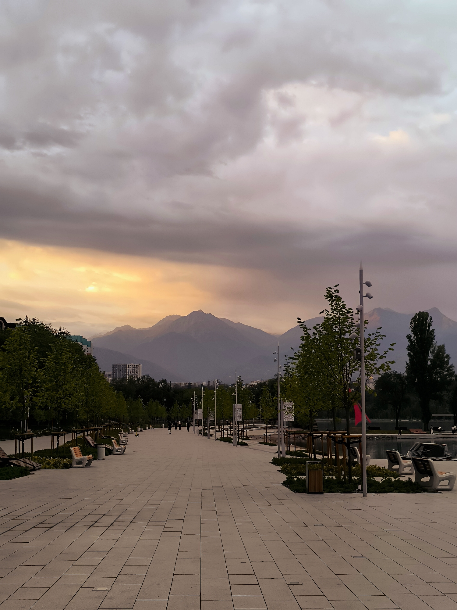 A paved walkway with benches and trees lines the left side of the image. In the distance, mountains are visible, partially obscured by clouds. The sky is overcast with a band of golden light breaking through near the horizon. People are walking in the distance on the walkway. The scene appears to be a park or public space in Almaty, Kazakhstan.