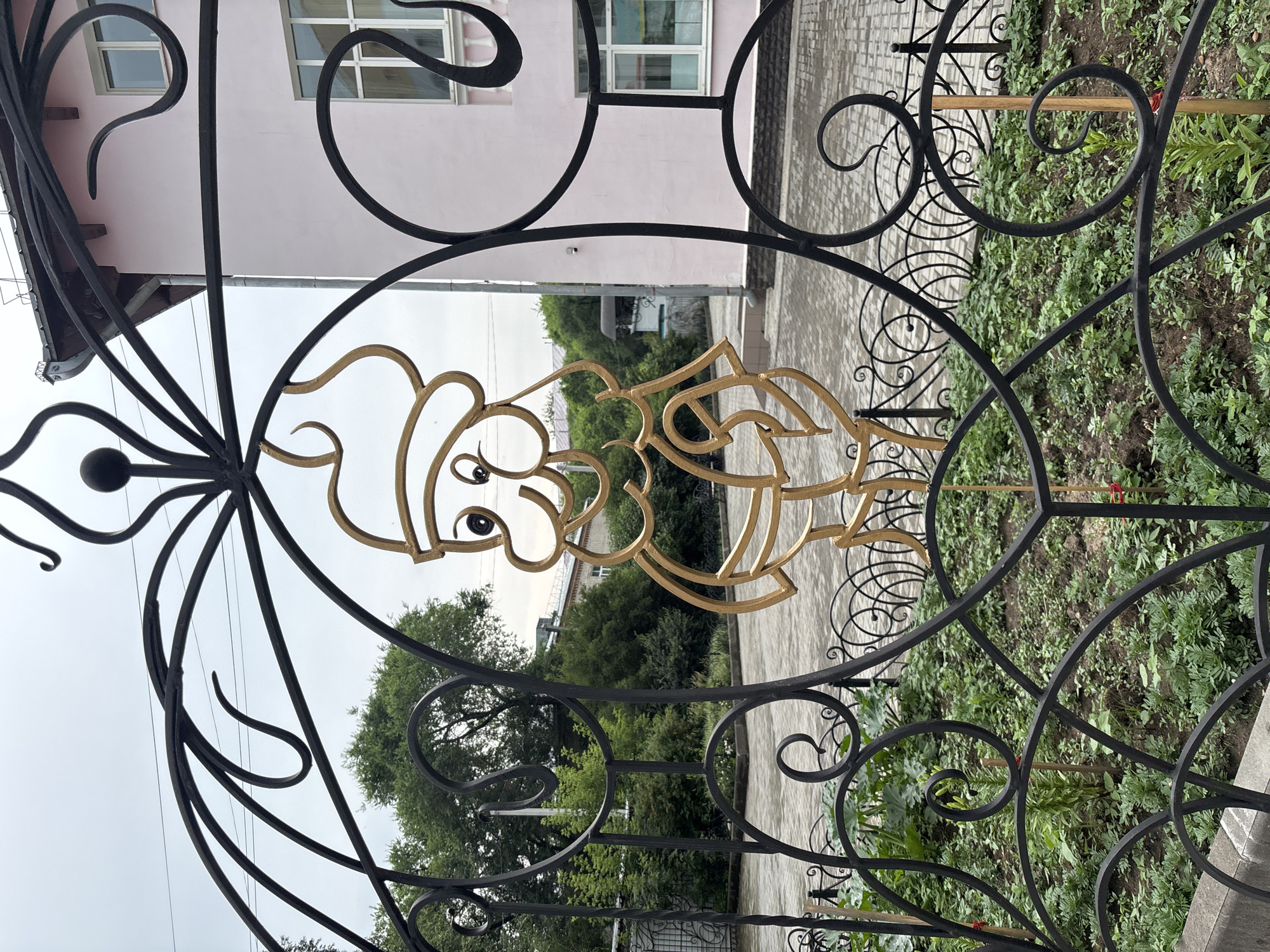 An ornate black metal gate with a gold cartoon character figure is in the foreground of the image. The character appears to be a stylized depiction of a figure wearing a hat and holding something. Behind the gate, a paved area with brick paving stones is visible, with greenery and trees beyond. To the right of the paved area is a light pink building with white-framed windows. The sky is overcast and gray. This gate is located in Blagoveshchensk, Russia, specifically at the entrance to the Blagoveshchensk State Pedagogical University.