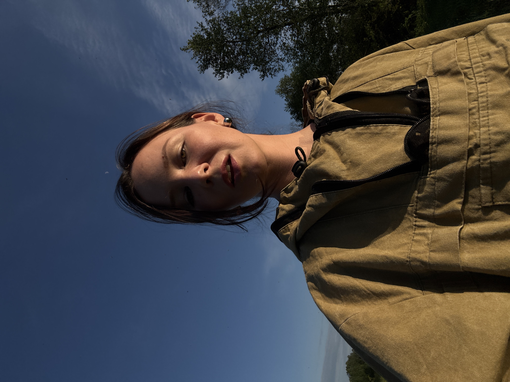 A low-angle, close-up shot depicts a woman outdoors. She has brown hair and is wearing a tan jacket. The sky is visible in the background, with trees at the top of the frame. The location appears to be outdoors and may be a park in Egorii, Russia given the sky and tree foliage.
