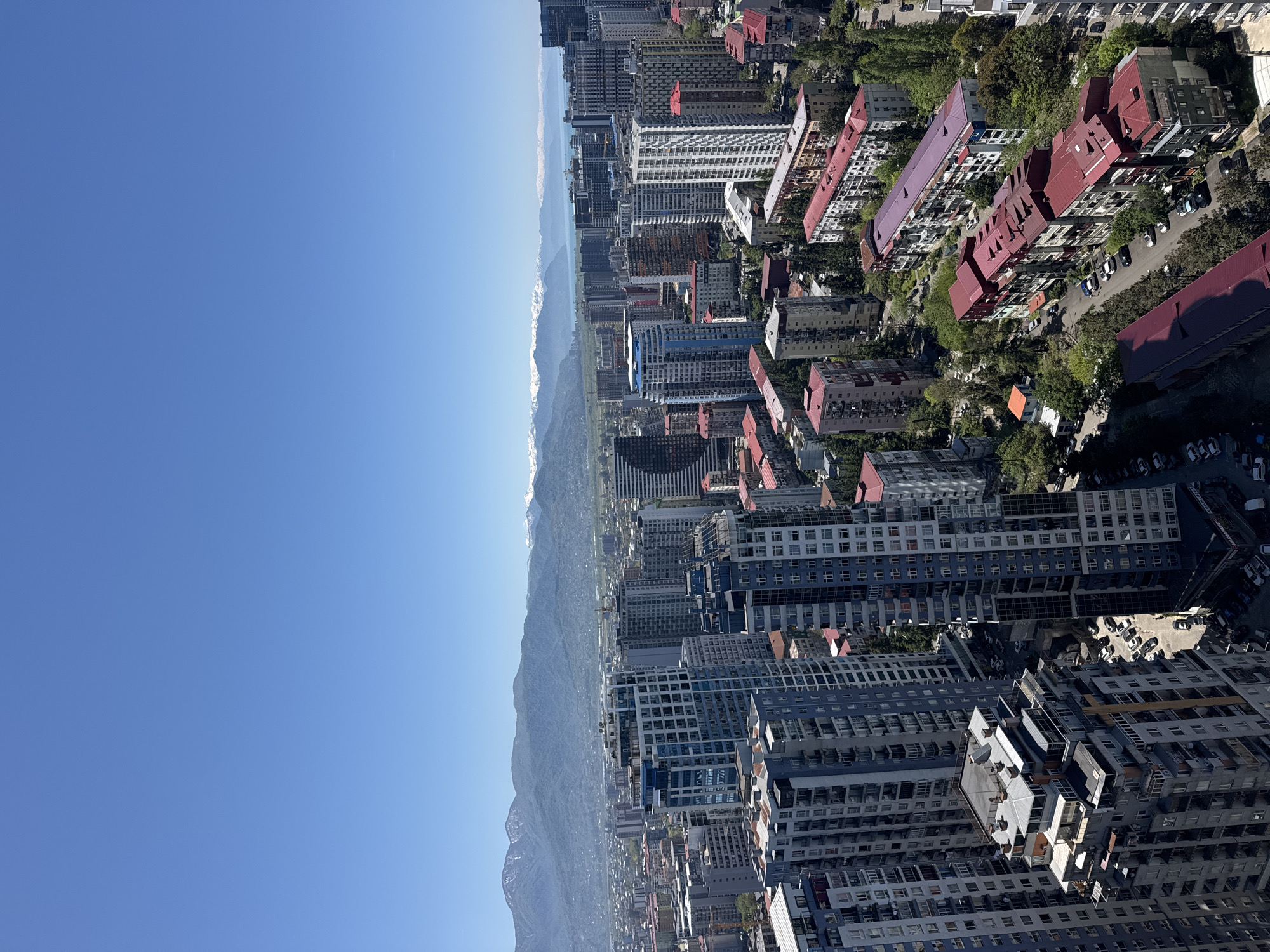 The image shows a cityscape of Batumi, Georgia, with a clear blue sky to the left and a mountain range dividing the sky from the urban area. The city's architecture is a mix of high-rise apartment buildings, some with a bluish-gray color scheme, interspersed with older structures featuring red roofs. Trees provide some greenery among the buildings. The mountains are visible in the distance with some snow-capped peaks. Roads with parked cars are seen among the buildings.