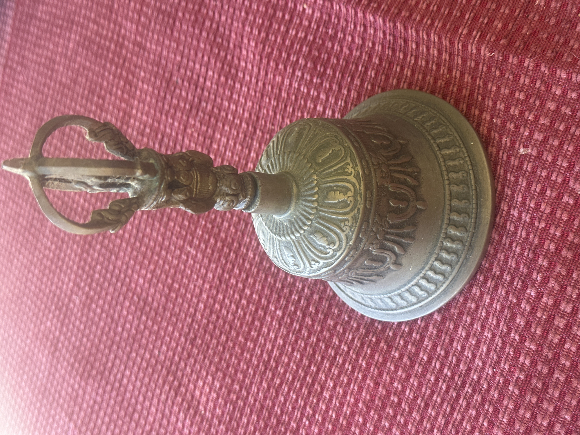 A close-up, eye-level shot shows a bronze ritual bell with intricate carvings sitting on a textured red fabric surface. The bell has a flared rim and a decorative band around its body. Atop the bell is a vajra, a symbolic scepter in Hinduism and Buddhism. The fabric has a repeating diamond-like pattern. The lighting casts subtle shadows on the bell and fabric, highlighting the textures. The background is a blurred expanse of the red fabric. There are no people visible, and the scene appears to be set indoors, on a flat surface. No specific location within Thora, Australia, can be determined from the visual information provided.