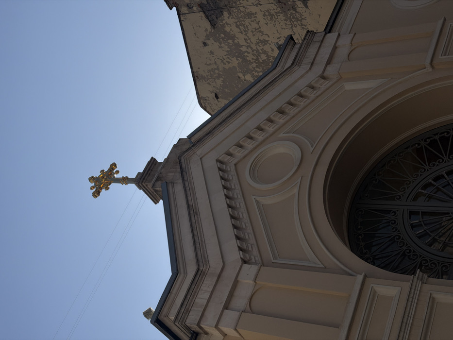 The image depicts an upward-angled view of the upper facade and roofline of the Armenian Cathedral of the Holy Transfiguration (Surb Amenaprkich Vank) in Moscow, Russia. The central subject is the cathedral's light beige, neoclassical stone or stucco facade, detailed with white trim, classical cornices, dentil-like moldings, rectangular recessed panels, and circular decorative elements. Below these details, a dark, ornate wrought iron arched double door is partially visible. Centrally positioned atop a stone plinth at the highest point of the facade, a multi-layered, ornate gilded cross, characteristic of Armenian Apostolic architecture, stands prominently against a clear, light blue sky. Two thin, dark electrical wires stretch horizontally across the sky in the upper left. To the right, a portion of a contrasting, darker, dilapidated building with peeling, yellowish-brown plaster is visible. No individuals are present in the frame.