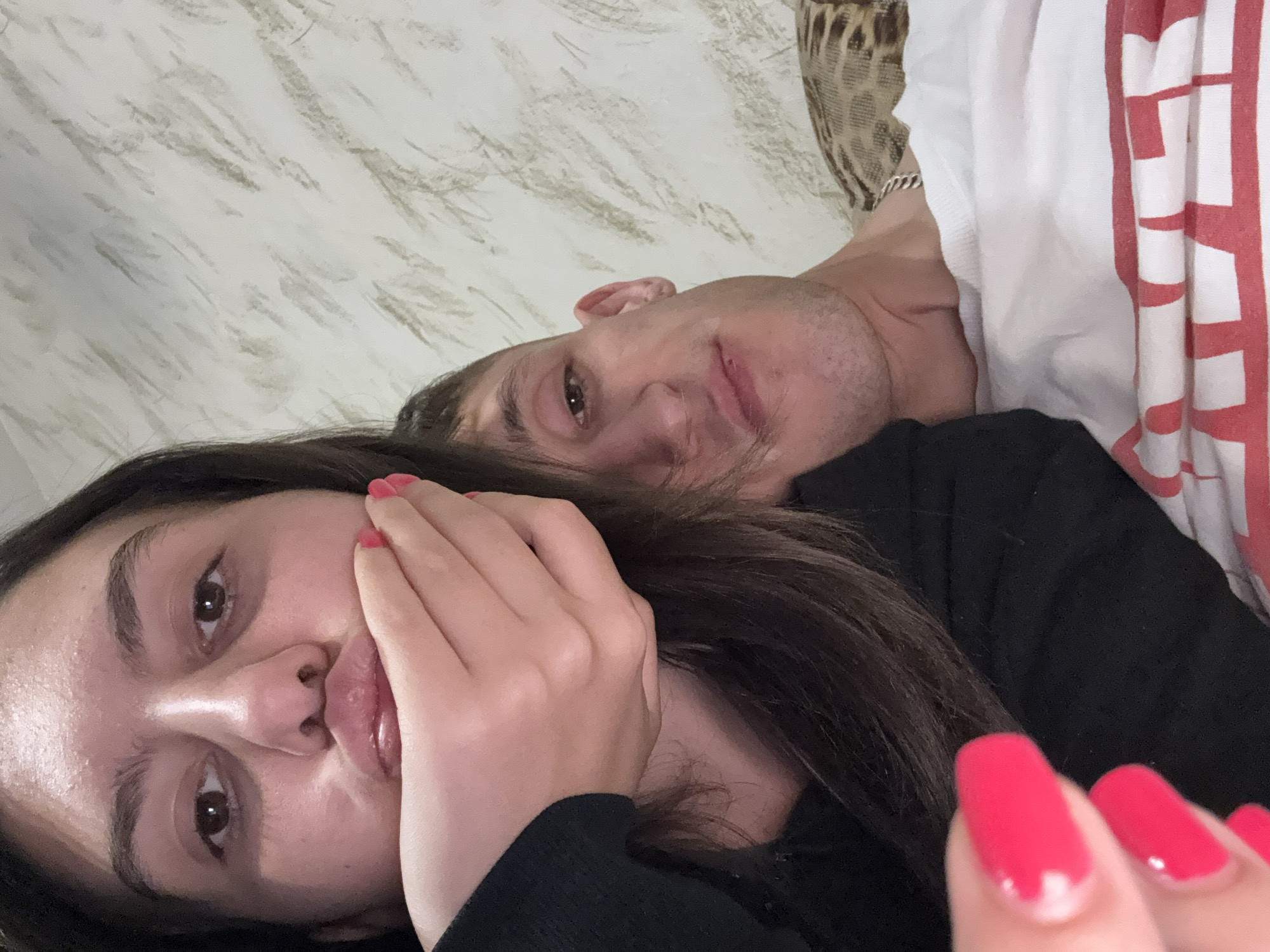 A close-up shot depicts a woman and a man lying next to each other. The woman is in the foreground with dark hair, a black top, and red nail polish, partially covering her mouth with her hand. Behind her is a man with short dark hair wearing a white and red shirt. The background consists of a textured wall and a leopard print fabric. There is no indication of the location being Stara Zagora, Bulgaria.