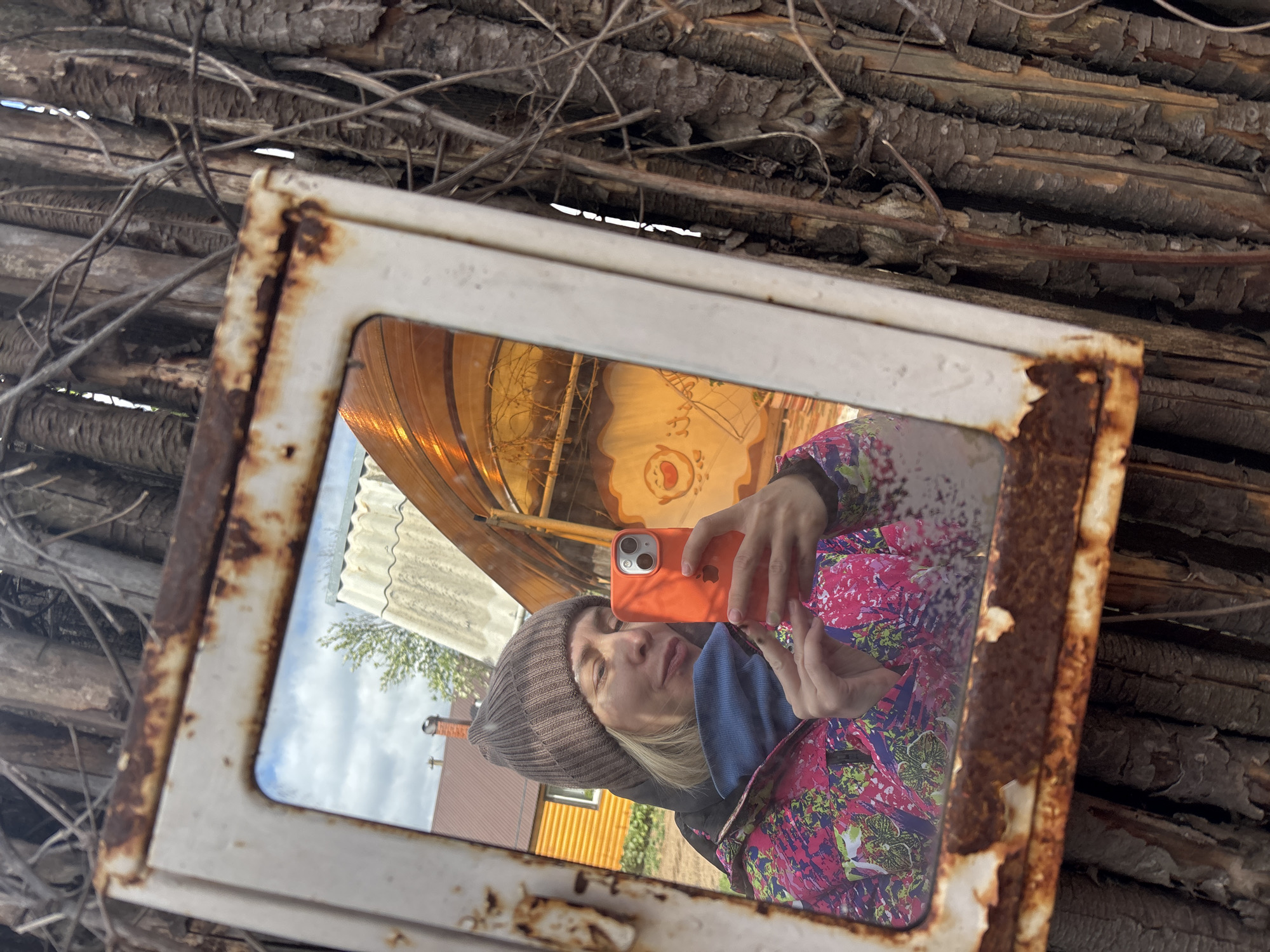A selfie is taken in a rusty, weathered metal-framed mirror. The mirror reflects a woman in a brown knit hat, blue scarf, and bright floral jacket, holding an orange iPhone. Behind her reflection are a house with a yellow exterior and a glimpse of trees and sky. The mirror is set against a backdrop of stacked, weathered logs and twigs. The location is likely Ваньки, Russia.