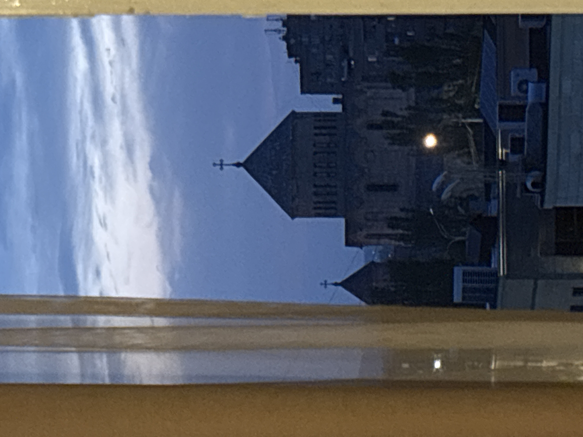 The image is a view from a window in Yerevan, Armenia, showing the St. Anna Church. The church is built of dark stone. A cross is affixed to the top of the tower. The sky behind the church is cloudy. Other buildings are visible in the background. The window frame is in the foreground, slightly blurred.