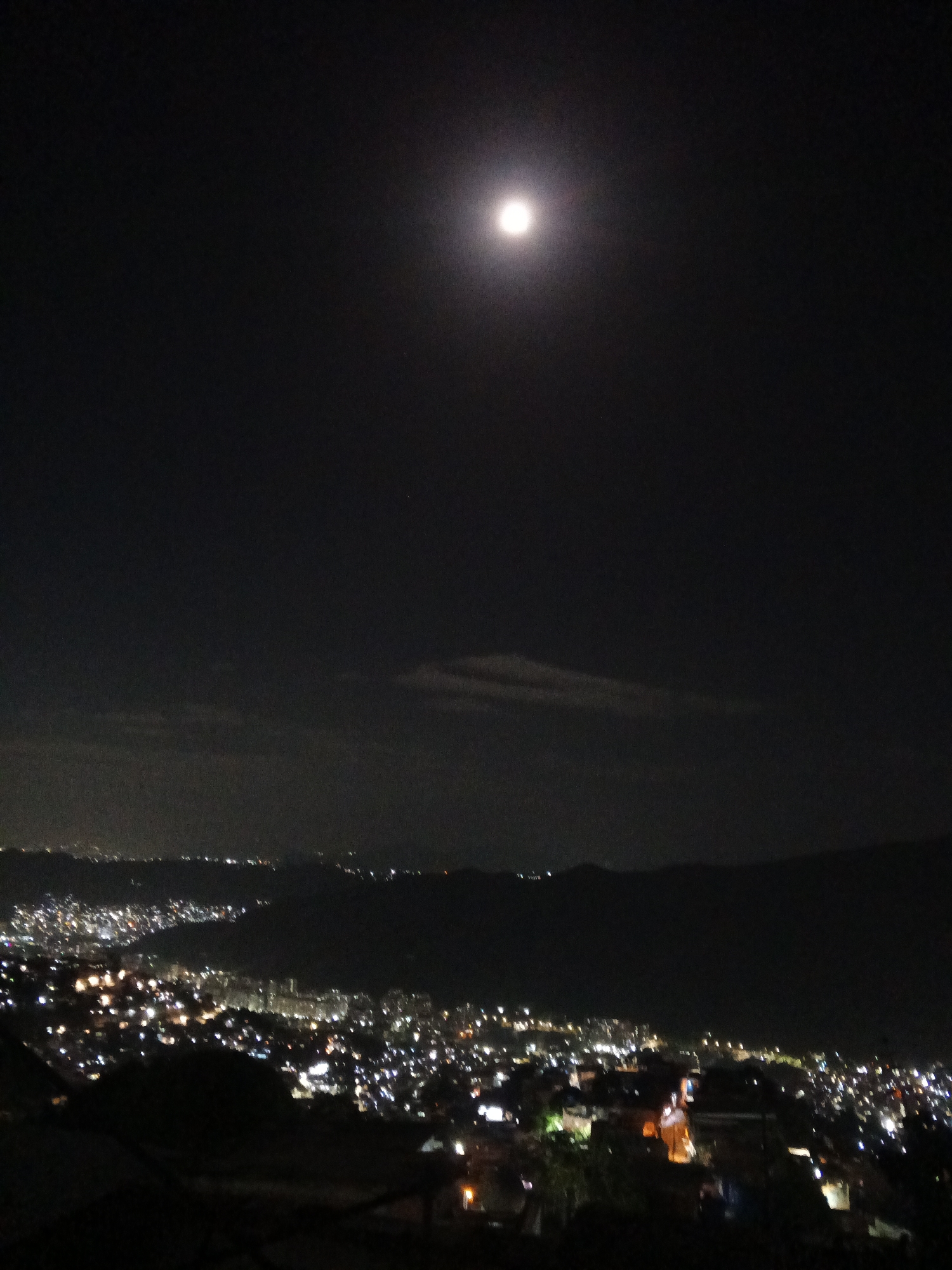 The image depicts a nighttime view of a city, likely Caracas, Venezuela, under a full moon. The city is spread out across a valley, with numerous lights illuminating buildings and streets. Mountain ranges form a backdrop. The sky is dark, with some cloud cover.