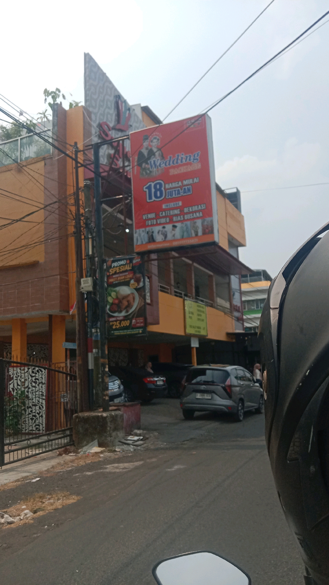 In Bekasi, Indonesia, a street scene unfolds. A prominent orange building with a peaked roof dominates the left side of the frame. On its facade, a large, red billboard advertises "Wedding Package" with an offer starting at "18 JUTA-AN" (18 Million Rupiah). Below this, it lists inclusions: VENUE, CATERING, DECORASI, FOTO VIDEO, RIAS BUSANA. Another smaller, dark banner hangs vertically, promoting "PROMO SPESIAL!" for "PAKET AYAM LEZAT" (delicious chicken package) for "Rp25.000".
In the foreground, the scene is captured from the perspective of someone on a motorcycle, with a portion of the helmet and mirror visible at the bottom and right edge of the frame respectively. The street is asphalt, showing signs of wear and tear, with scattered debris near a concrete structure on the left. Several cars are parked or driving on the street, including a dark grey hatchback in the middle ground and a darker vehicle behind it. Power lines crisscross the sky above. The overall atmosphere is that of a bustling urban environment.