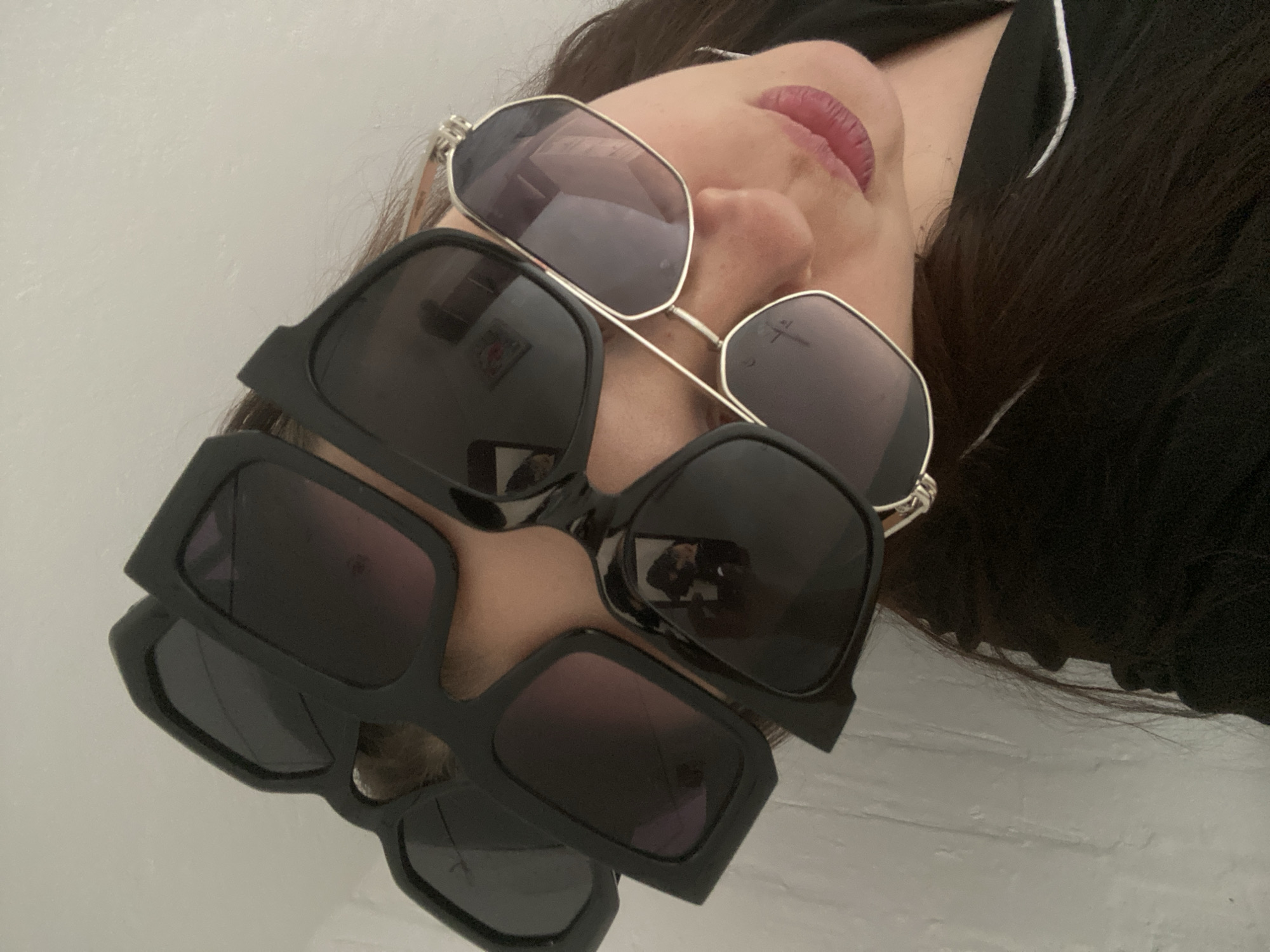 A woman lies on her back wearing a black top and three pairs of sunglasses. The uppermost pair is silver-rimmed and has light purple lenses. The next two pairs are black, rectangular, and stacked on top of each other. Her lips are visible and appear to be red. The background is a light-colored, possibly white, wall. The location is not discernible from the image.