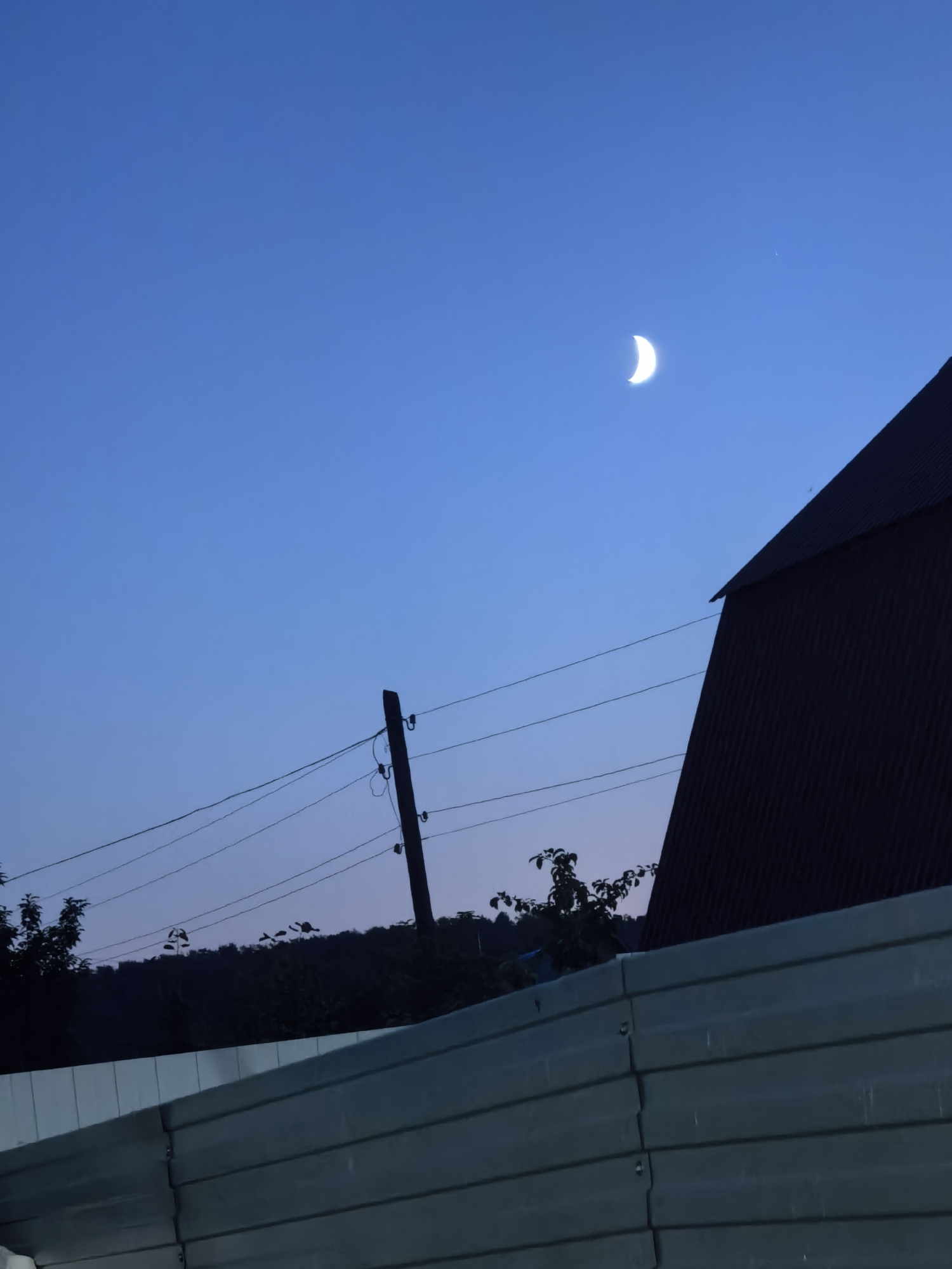 An outdoor, eye-level, full shot captures a darkening sky with a sliver of a crescent moon visible in the upper right quadrant. In the foreground, a muted teal corrugated metal fence runs horizontally across the bottom of the frame. Above the fence, a dark silhouette of trees and a tree line create a textured horizon. A dark, angular roofline of a building juts into the frame from the upper right. A wooden utility pole with wires stretches diagonally from the middle left to the mid-right of the frame, silhouetted against the twilight sky. The scene appears to be in a rural or suburban setting.