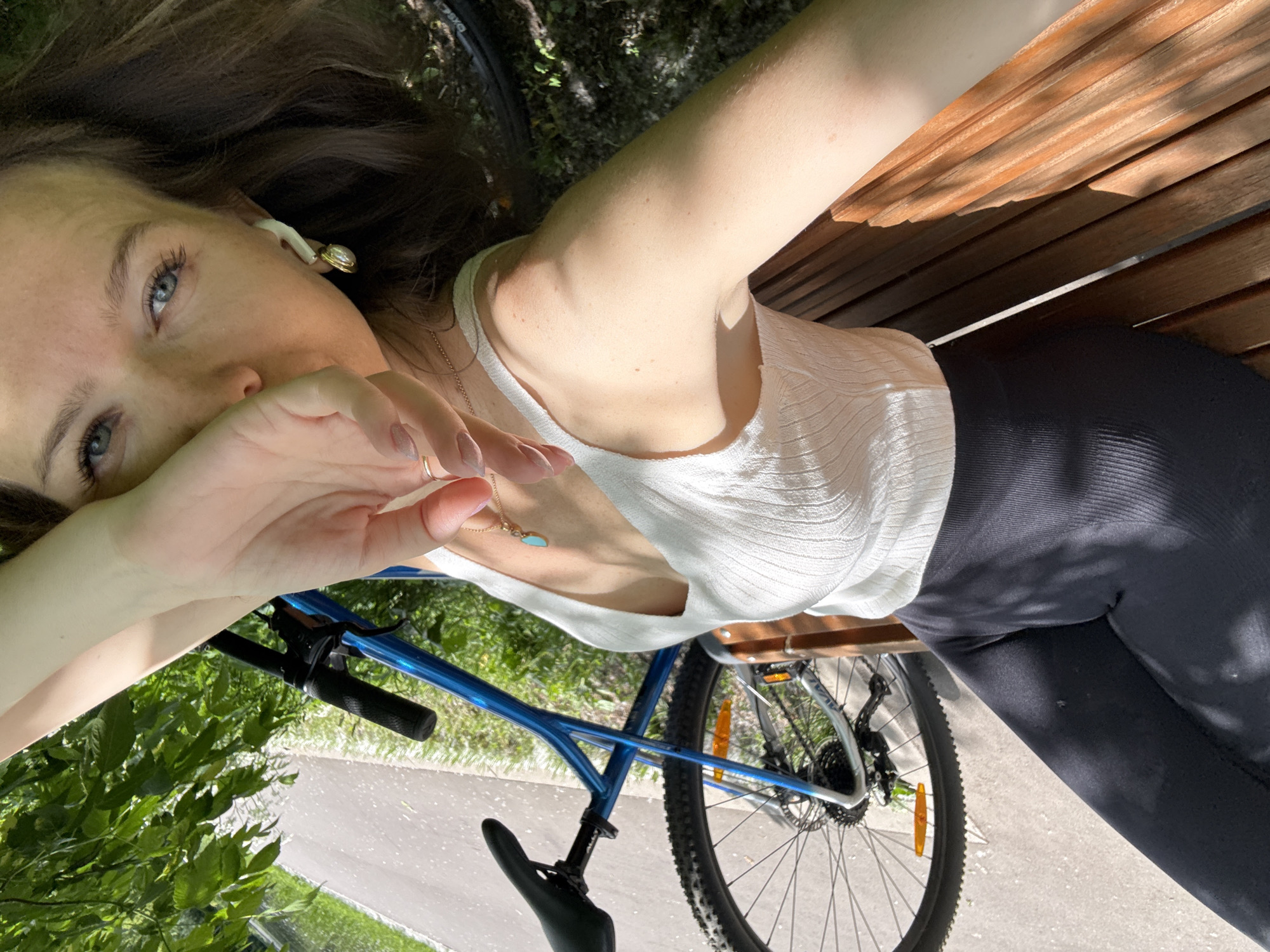 The image presents a high-angle, tilted selfie of a young woman resting outdoors, likely on a wooden park bench, in Moscow, Russia. Her dark brown hair spreads out, and her striking blue eyes gaze towards the camera, with her left hand partially covering her mouth and nose. She wears a white, ribbed sleeveless top, dark form-fitting leggings, and a delicate gold chain necklace with a small turquoise pendant. A unique gold earring with a white element is visible on her right ear, along with a white earbud. A gold ring adorns her left ring finger.
A blue bicycle is a prominent feature in the foreground and midground, suggesting a break during a ride. Its handlebars, frame, seat, and robust-treaded wheels are clearly visible. The setting is outdoors, characterized by bright sunlight casting shadows, lush green foliage, and a paved path, indicating a park or a recreational area. The wooden slats of the bench she is on are visible behind her. The atmosphere is relaxed and casual, capturing a moment of repose on a sunny day. No visible text appears in the image.