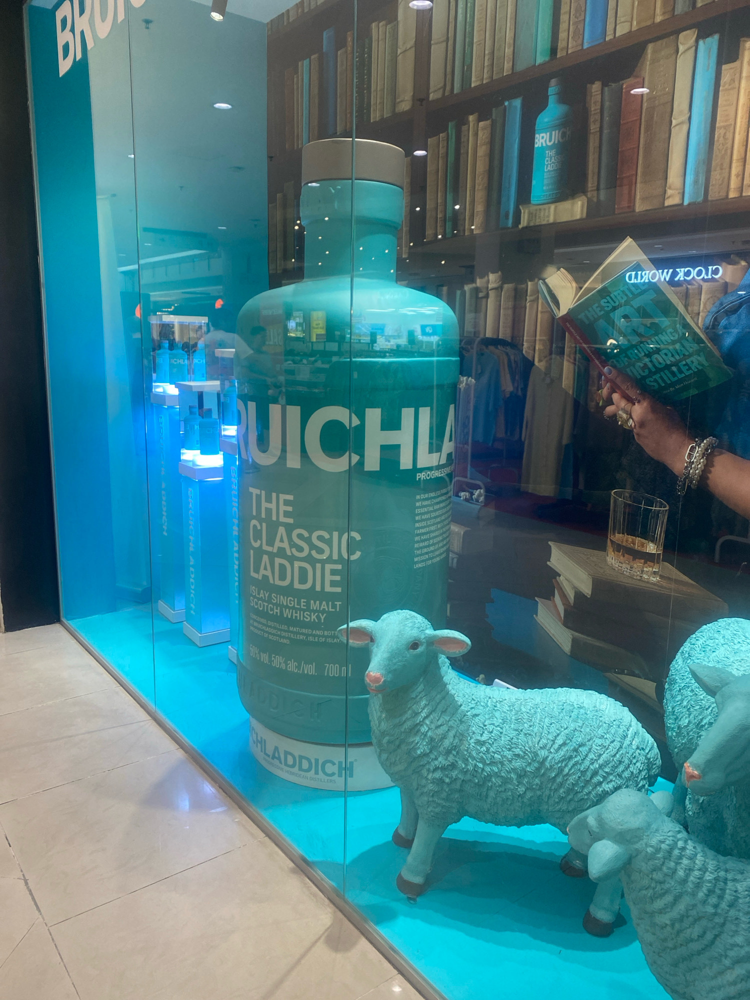 The image captures a store window display with a turquoise theme. Dominating the center is a giant replica of a Bruichladdich "The Classic Laddie" whisky bottle. In front of the bottle, several light blue, stylized sheep figurines are positioned. The background features a bookshelf filled with books, a person's hand holding an open book, and a glass of amber liquid on a stack of books. To the left, a vertical sign displays the word "BRUICHLADDICH" in large white letters against a turquoise background. Multiple smaller Bruichladdich bottles are displayed on illuminated stands to the far left. The flooring is tiled. The scene appears to be in a shopping mall. Based on the style of the store and common luxury brand presence, this display is likely located in a high-end shopping district in Kuala Lumpur, Malaysia, such as Suria KLCC.