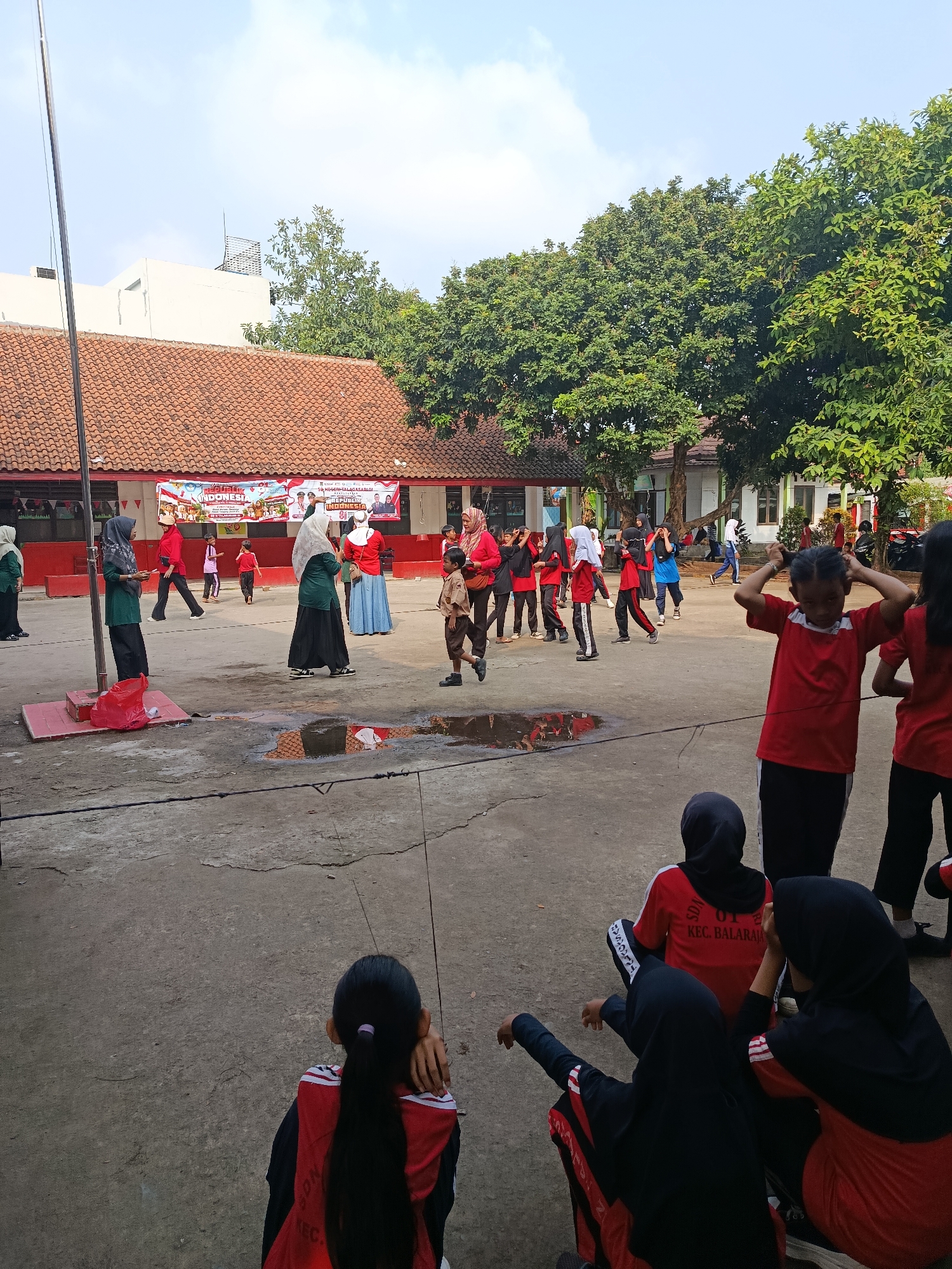 In an outdoor schoolyard in Balaraja, Indonesia, a group of students and adults are gathered. The scene is set on a paved surface with some puddles of water. A large red and white banner with Indonesian text and images of political figures is prominently displayed on the side of a building. The building appears to be a school, with red tiled roofs and white walls. Trees with lush green foliage are visible in the background.
A group of individuals, mostly wearing red shirts and some with headscarves, are actively participating in an event. Some are standing and looking towards the banner, while others appear to be in motion. Several children are also present, some in uniforms. A flagpole stands to the left of the frame. The sky is partly cloudy and appears to be daytime. The overall atmosphere suggests a community event or a school activity taking place.