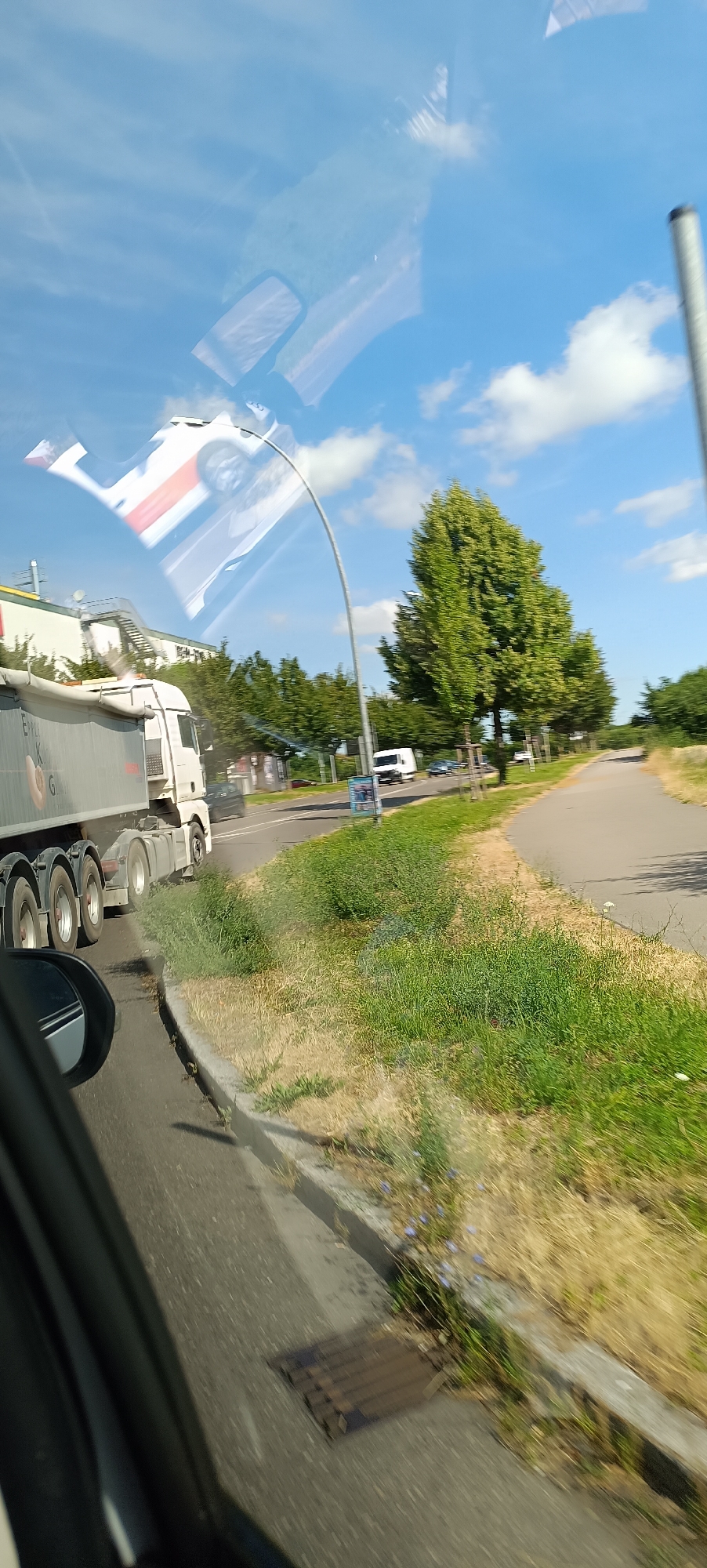 A reflective surface, likely a car windshield, distorts the view of a street scene in Fellbach, Germany. A large white truck with a gray trailer is on the road. A white panel van and other vehicles are visible further down the road. Trees line the right side of the road, with a sidewalk or bike path adjacent to them. The sky is blue with scattered white clouds. The reflection on the windshield shows a distorted image of the sky and possibly parts of a car interior.