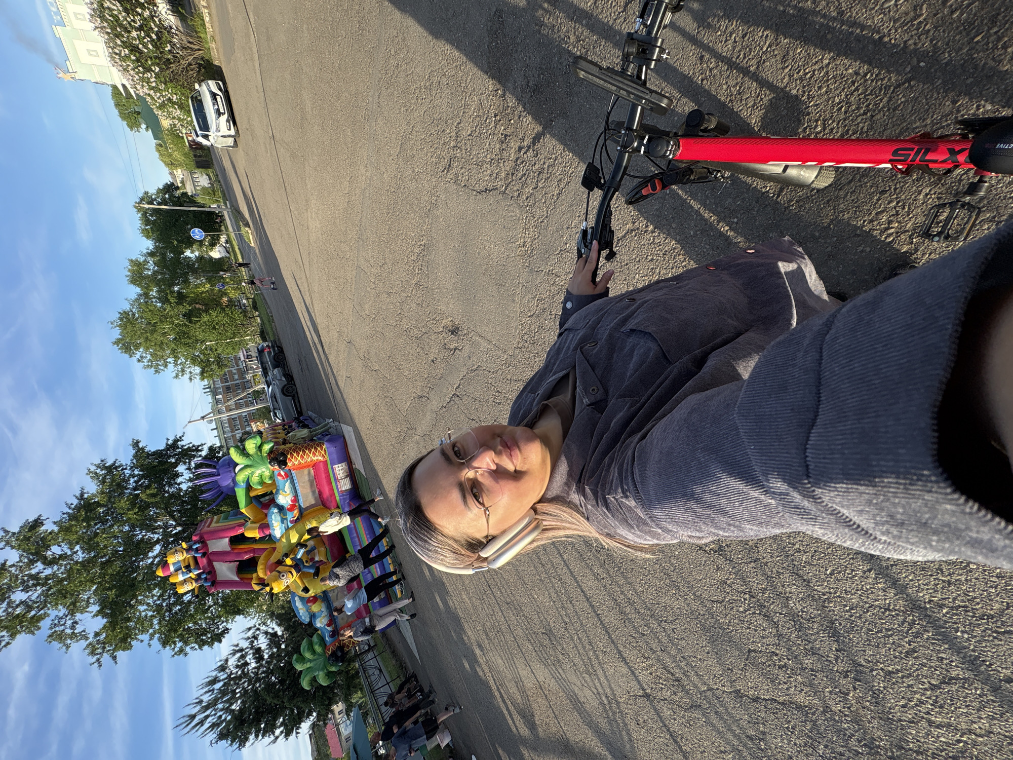 The image shows a person, likely a woman, taking a selfie while on a bicycle. The bicycle has a red frame and black handlebars. The person is wearing glasses and headphones, and a grey shirt. The background includes a road, trees, a bouncy castle, and people gathered near it. In the distance, there are buildings and a car. Based on visual clues, the location might be a park or public space within a city; however, I am unable to verify the location in Birobidzhan, Russia.
