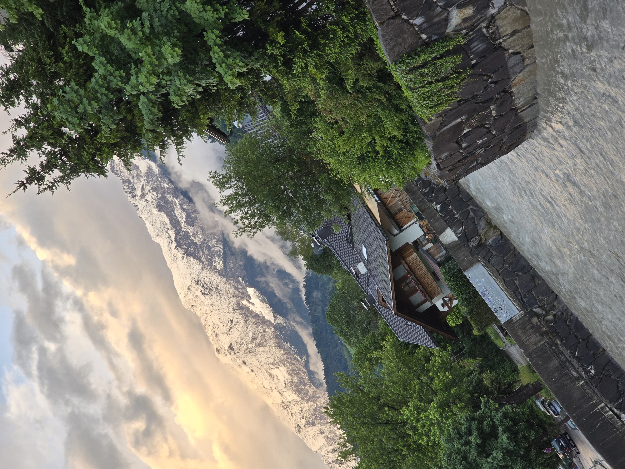 The image depicts a scene in Garmisch-Partenkirchen, Germany, characterized by a powerful river in the foreground, flowing between stone retaining walls covered in ivy. The river's surface is turbulent, suggesting a strong current. Beyond the river, a village is nestled amongst lush green trees. Traditional Bavarian-style houses with dark tiled roofs and wooden balconies are visible. In the background, a majestic snow-capped mountain range dominates the horizon. The sky is a dramatic mix of warm-toned clouds, illuminated by what appears to be the setting or rising sun, and cooler, grayer cloud formations. The overall impression is one of natural beauty, with the imposing mountain landscape contrasting with the human-made structures and the lively river.