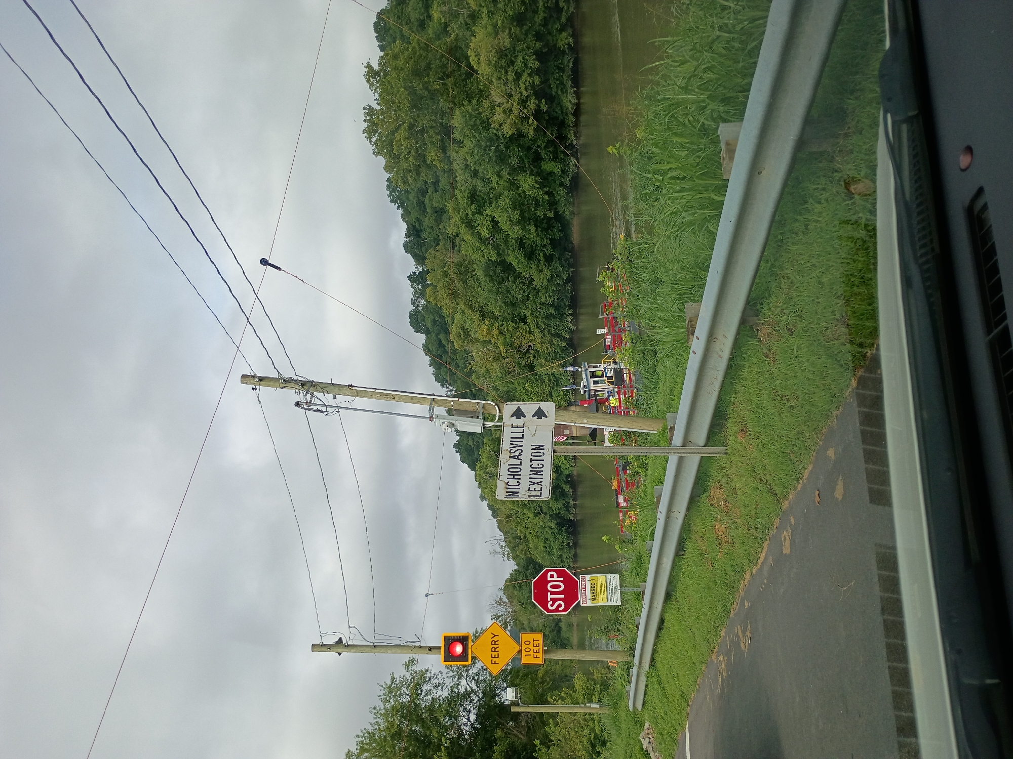 The image shows a road leading to a ferry crossing on a cloudy day. A red traffic light is illuminated, indicating that the ferry is not currently boarding. A yellow diamond-shaped sign indicates "FERRY 100 FEET." A red octagonal STOP sign is also visible, along with directional signs pointing to "NICHOLASVILLE" and "LEXINGTON." A barge or ferry is docked on the other side of the river, and people in yellow vests are visible on the barge. The river is flanked by lush green trees and vegetation. Power lines crisscross the sky. The foreground shows the hood and windshield of a vehicle, suggesting the photo was taken from a car. The scene appears to be a ferry crossing in a rural area.