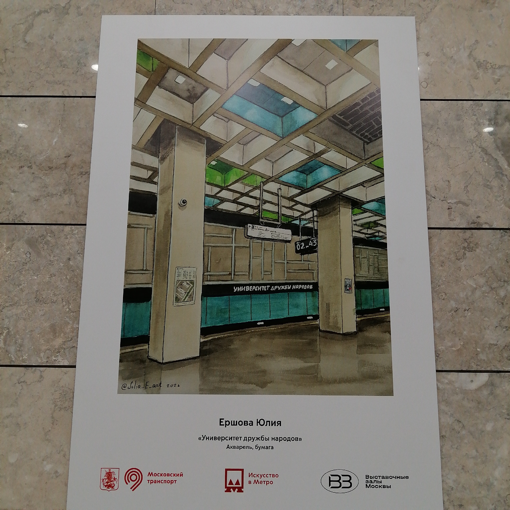 The image shows a watercolor painting of the "Universitet Druzhby Narodov" (Peoples' Friendship University) metro station in Moscow, Russia. The painting depicts the interior of the station, including the platform area with its distinctive architectural features, such as the columns, the ceiling structure with rectangular panels, and the platform wall with the station name in Cyrillic. A sign with the numbers "02-43" is visible. The painting is displayed on a white poster, with the artist's name, "Ершова Юлия" (Ershova Yulia), the title of the artwork, and the medium used (watercolor on paper) written below the image. At the bottom of the poster, there are logos representing the "Moscow Transport," "Art in the Metro," and "Exhibition Halls of Moscow."