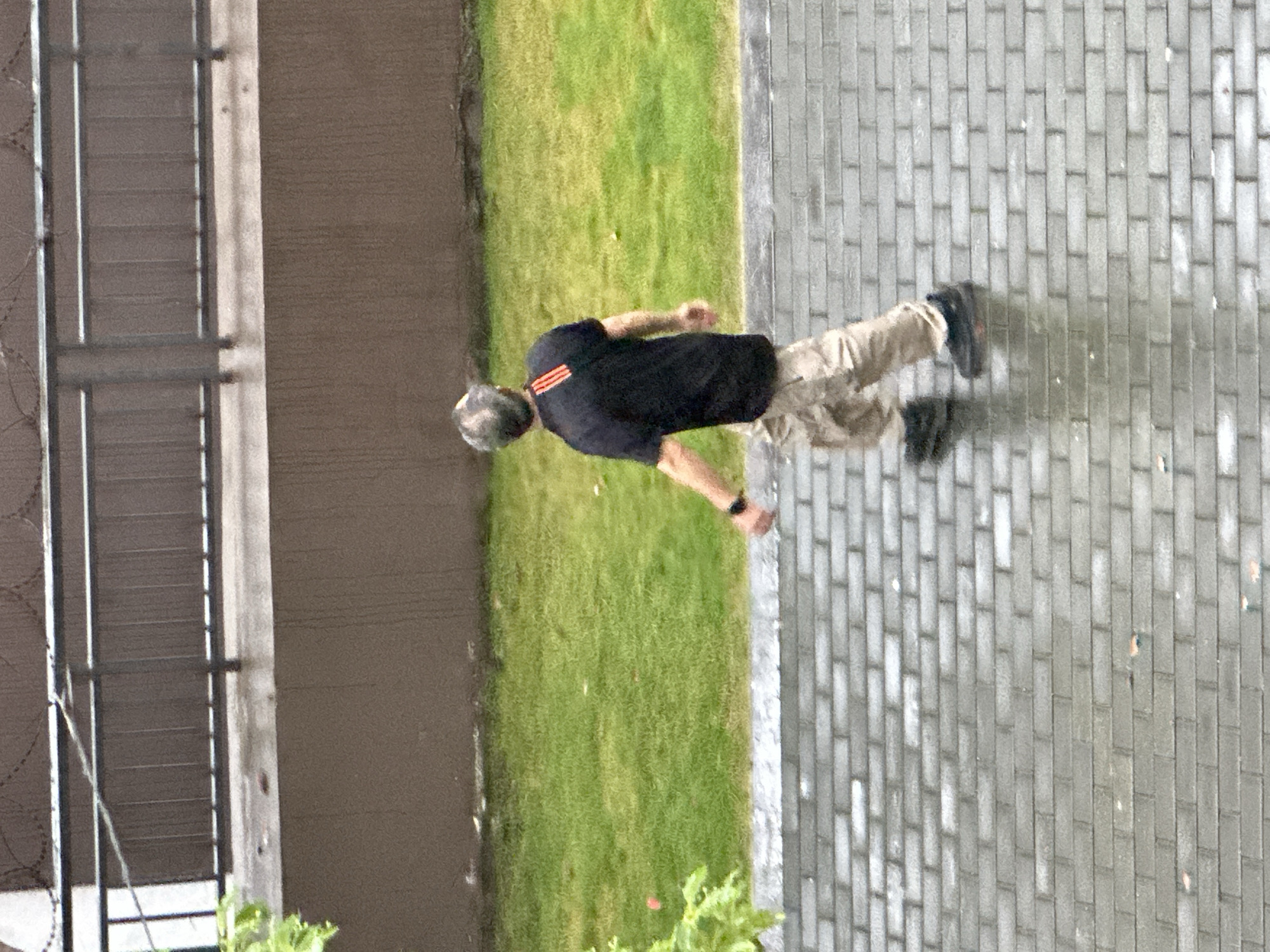 A person is walking on a paved pathway next to a grassy area. The person is wearing a black t-shirt with three orange stripes on the left sleeve and camouflage pants. They are also wearing black shoes. The pathway is made of gray bricks and appears to be wet, suggesting it may have rained recently or there is water on the ground. To the left of the pathway and grass is a building with a textured brown wall and a metal fence with sharp wire. The scene is likely outdoors in a developed area, possibly a park or a public walkway within the Chongqing High-tech Zone in China. The time of day is unclear, but the lighting suggests it is daytime. No text is visible in the image.