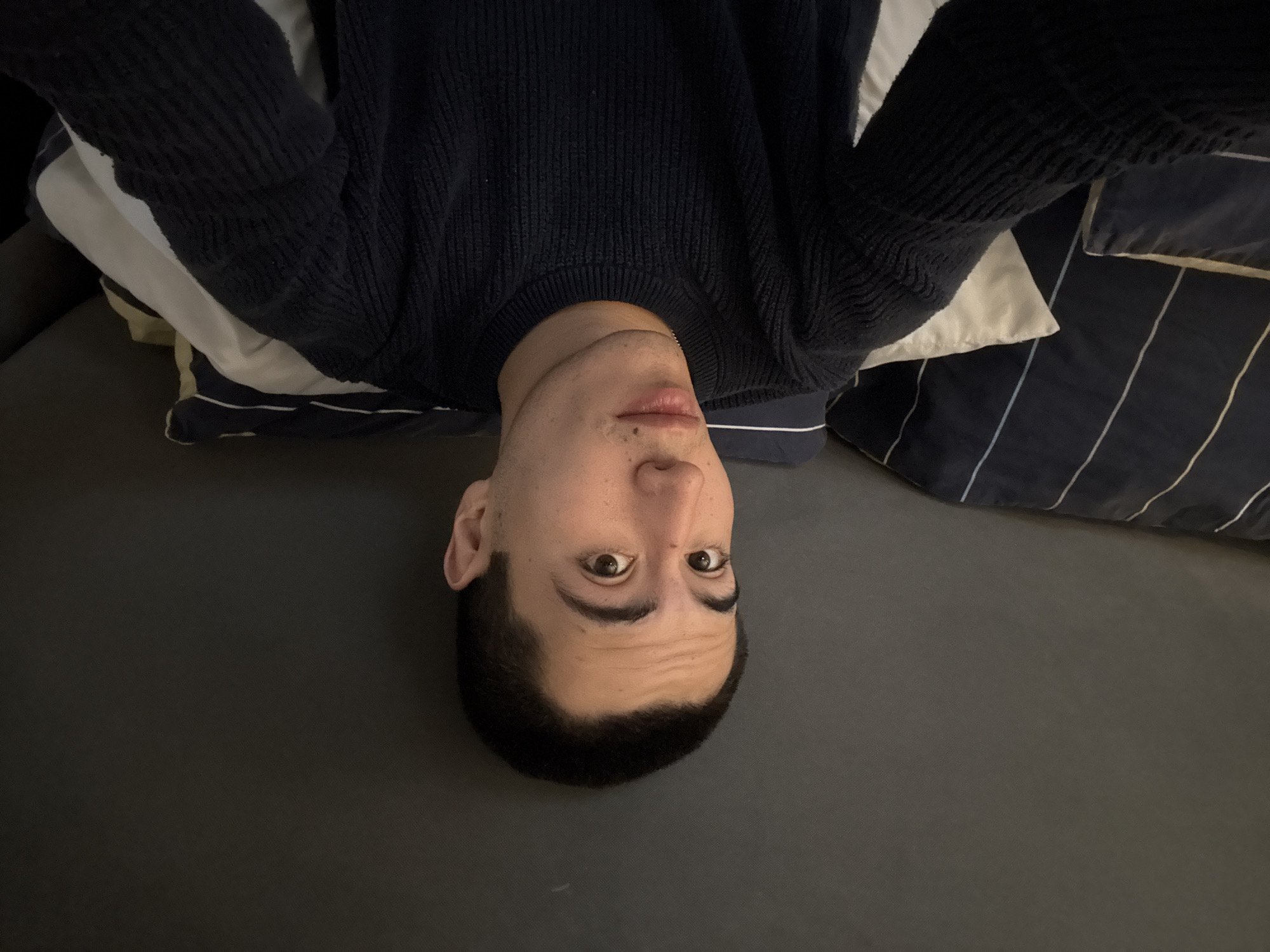 A man is lying on his back, upside down, on a dark gray surface that appears to be a bed. He is wearing a dark blue sweater. Pillows with white and blue patterned covers are behind his head. The location is unknown.