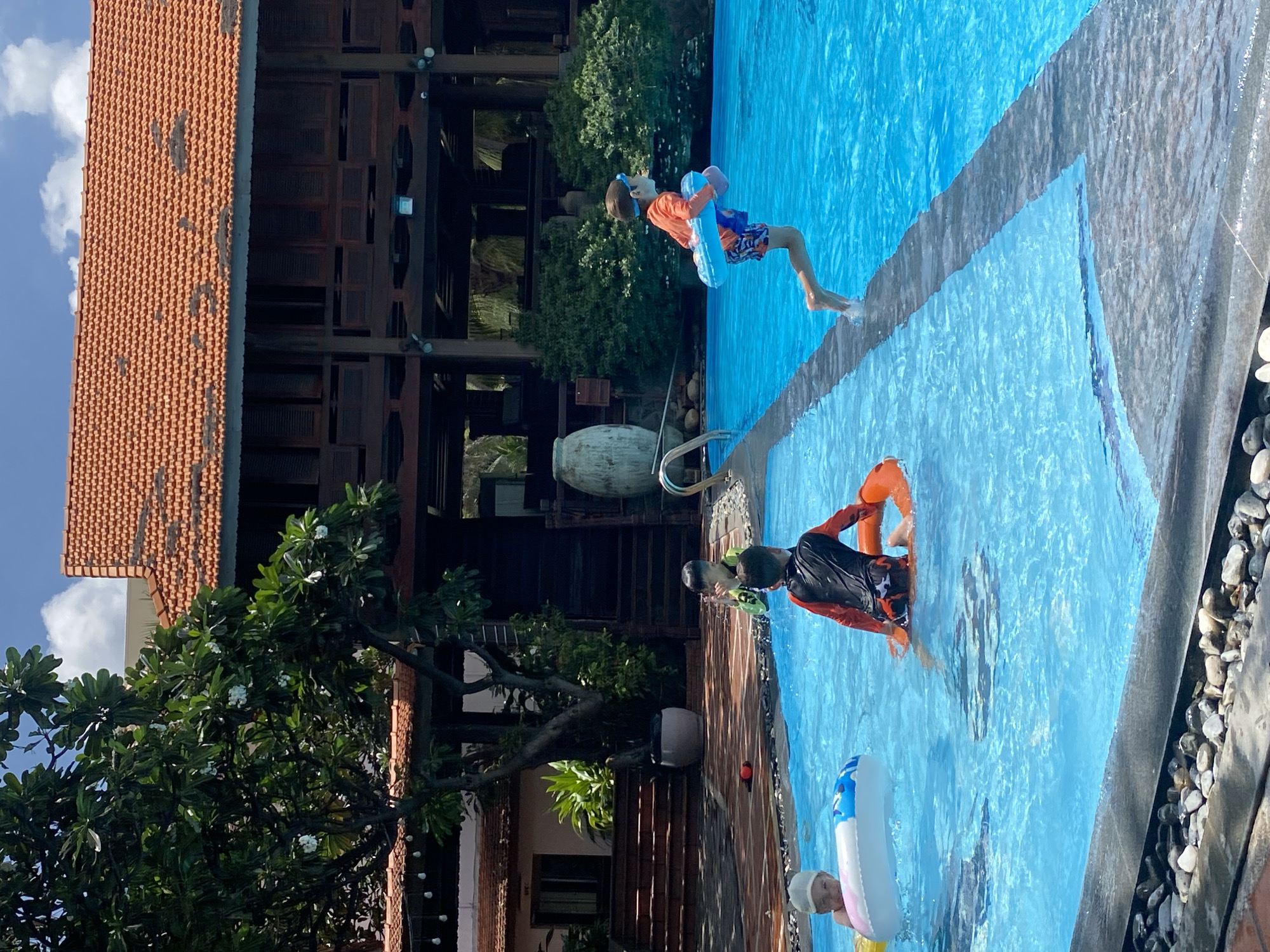The image depicts an outdoor swimming pool at a resort in Phu Thuy Ward, Vietnam. Several children are engaged in various water activities. In the foreground, a young child wearing a hat is in a blue and white inflatable ring. To the right, a child in a black and orange swimsuit is in an orange flotation device, appearing to be seated in the water. Further to the right, a child in an orange shirt and blue shorts is jumping into the pool, holding a blue inflatable ring. In the background, a building with a terracotta tile roof and dark wooden accents is visible, surrounded by lush greenery. The sky is bright blue with scattered white clouds. The pool water is a vibrant turquoise.