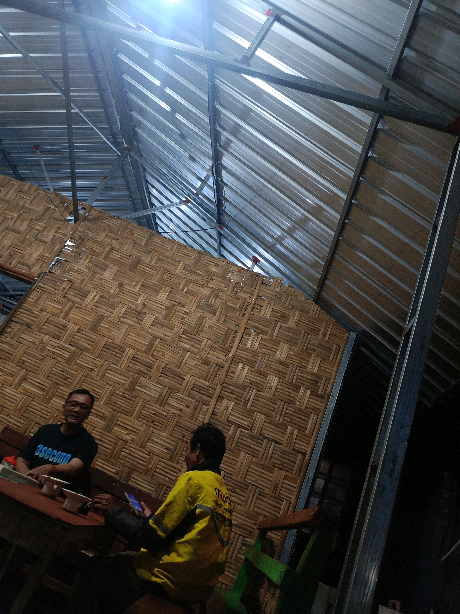 The image depicts an indoor scene, likely a cafe or restaurant, with a thatched roof constructed from woven bamboo. Two men are seated at a table in the foreground. One man, wearing a black t-shirt with "3second" written in blue, is facing the camera and smiling. The other man, wearing a yellow jacket with reflective stripes and the word "maxim" visible, is looking away from the camera and holding a smartphone. The environment suggests a casual gathering, possibly in Magetan, Indonesia, given the use of bamboo and the general aesthetic.
