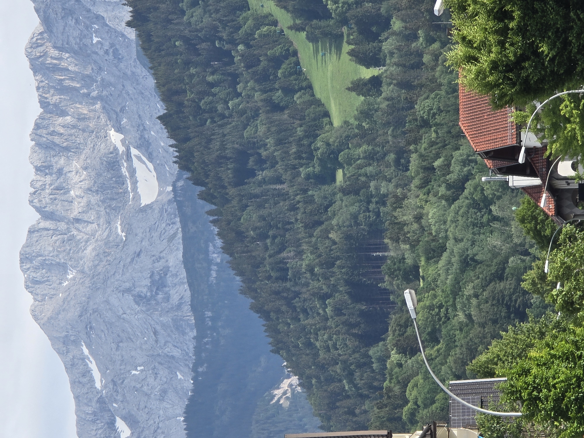 The image captures a mountainous landscape overlooking a town, likely in Garmisch-Partenkirchen, Germany, given the presence of alpine scenery. In the foreground, a streetlamp with a curved arm is visible, positioned to the left of the frame. Partially obscured structures of buildings, including a section of a dark-tiled roof and solar panels, are also present in the lower left and right portions of the image. Dense green foliage, consisting of trees and shrubs, dominates the mid-ground, creating a transition between the urban elements and the mountains.
Further into the mid-ground, the landscape ascends into a densely forested mountain slope. Patches of open green fields are interspersed within the forest, some of which appear to be maintained, possibly for recreational purposes. The texture of the forest is varied, with numerous coniferous trees forming a thick canopy.
The background is dominated by a vast, rugged mountain range. The peaks are sharp and rocky, with visible geological striations and some patches of snow, indicating a significant elevation. The mountain range exhibits layers of rock and scree, with less vegetation at higher altitudes. The sky is overcast and a pale gray, suggesting a cloudy or hazy day. No people are visible in the image. The overall impression is one of a natural landscape integrated with a human settlement, with the mountains serving as a prominent backdrop.