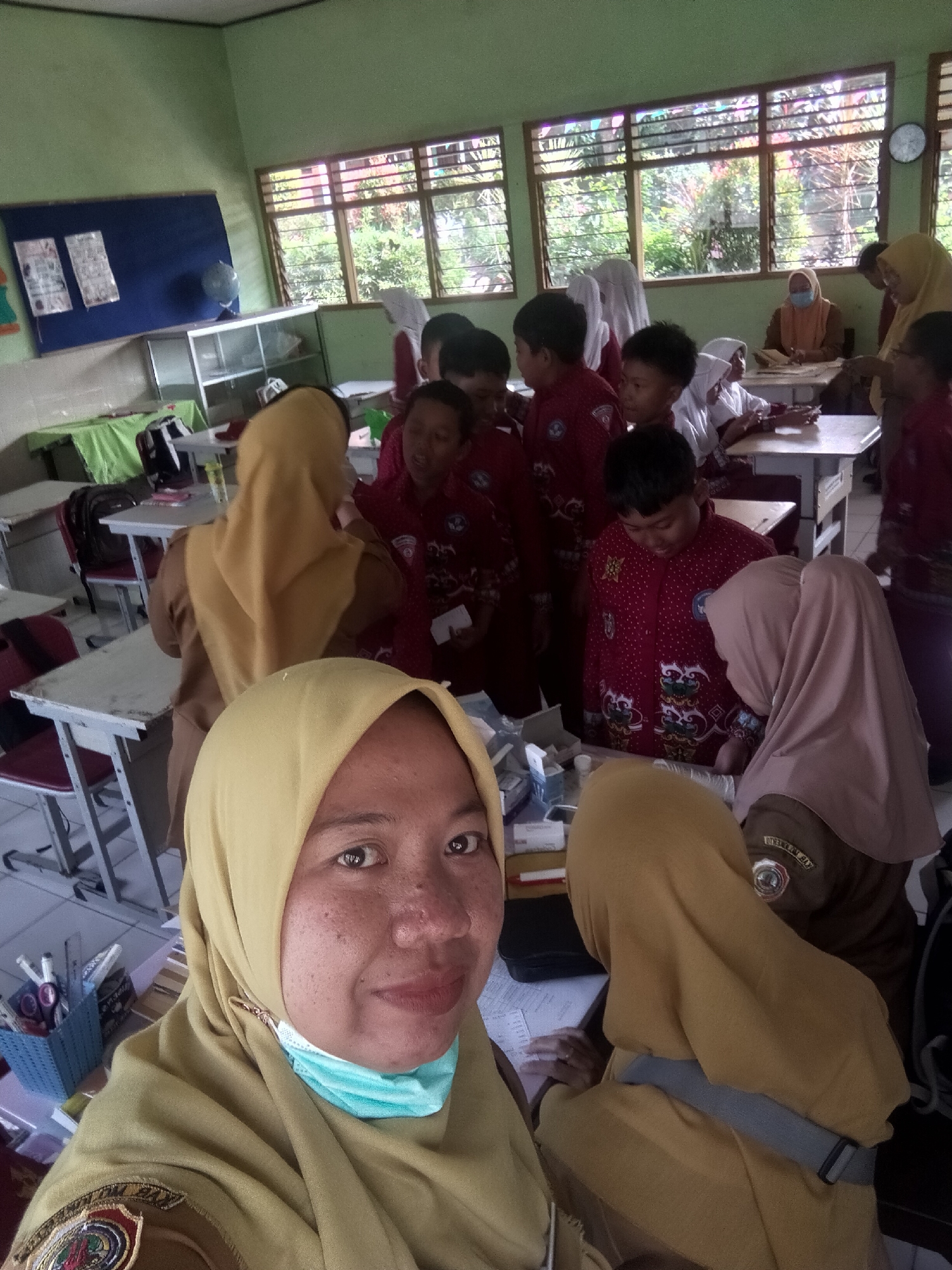 The image shows a group of people, mostly children wearing red uniforms and hijabs, in what appears to be a classroom. The room has green walls and windows with louvers, suggesting it is in a warm climate. Desks and chairs are visible, as well as a bulletin board with papers and a globe. A woman in a yellow hijab is in the foreground, taking a selfie. Another woman, also wearing a hijab, is seated at a desk with some children around her. The presence of medical supplies on a table suggests a possible health-related activity or demonstration. The scene is consistent with a school environment in Tretes, Indonesia.