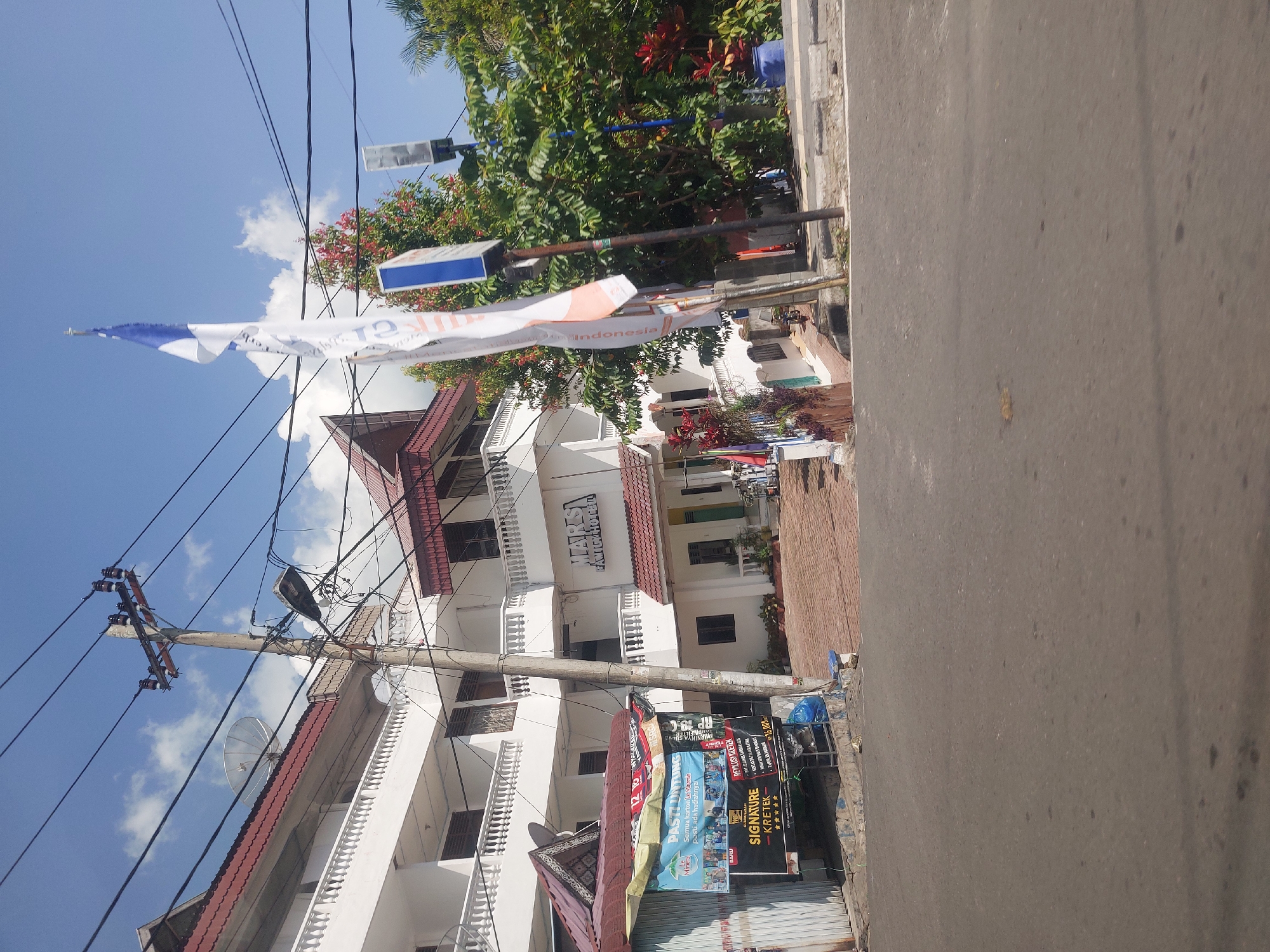The image shows a street scene in Tiga Raja, Indonesia, on a bright, sunny day. In the foreground, there is a paved road with some debris. To the left, a white building with multiple levels and balconies is visible. Adjacent to this building, a small, open-fronted kiosk or shop with a corrugated metal roof is present. This kiosk displays several colorful banners, one of which features text and images related to a cigarette brand and promotions.
Behind the kiosk and to the right of the white building is a larger, multi-story building with white walls and red-tiled roofs. A sign on this building reads "MARS FAMILY HOTEL." Flags are seen flying on poles near the hotel, including a white flag with blue accents. Electrical poles with numerous wires and a street lamp are also prominent features in the scene. Lush green trees and some red foliage are visible behind the hotel and along the right side of the image, with potted plants and a blue plastic container. The sky is blue with scattered white clouds. The overall impression is of a commercial area with lodging facilities and street-level businesses.