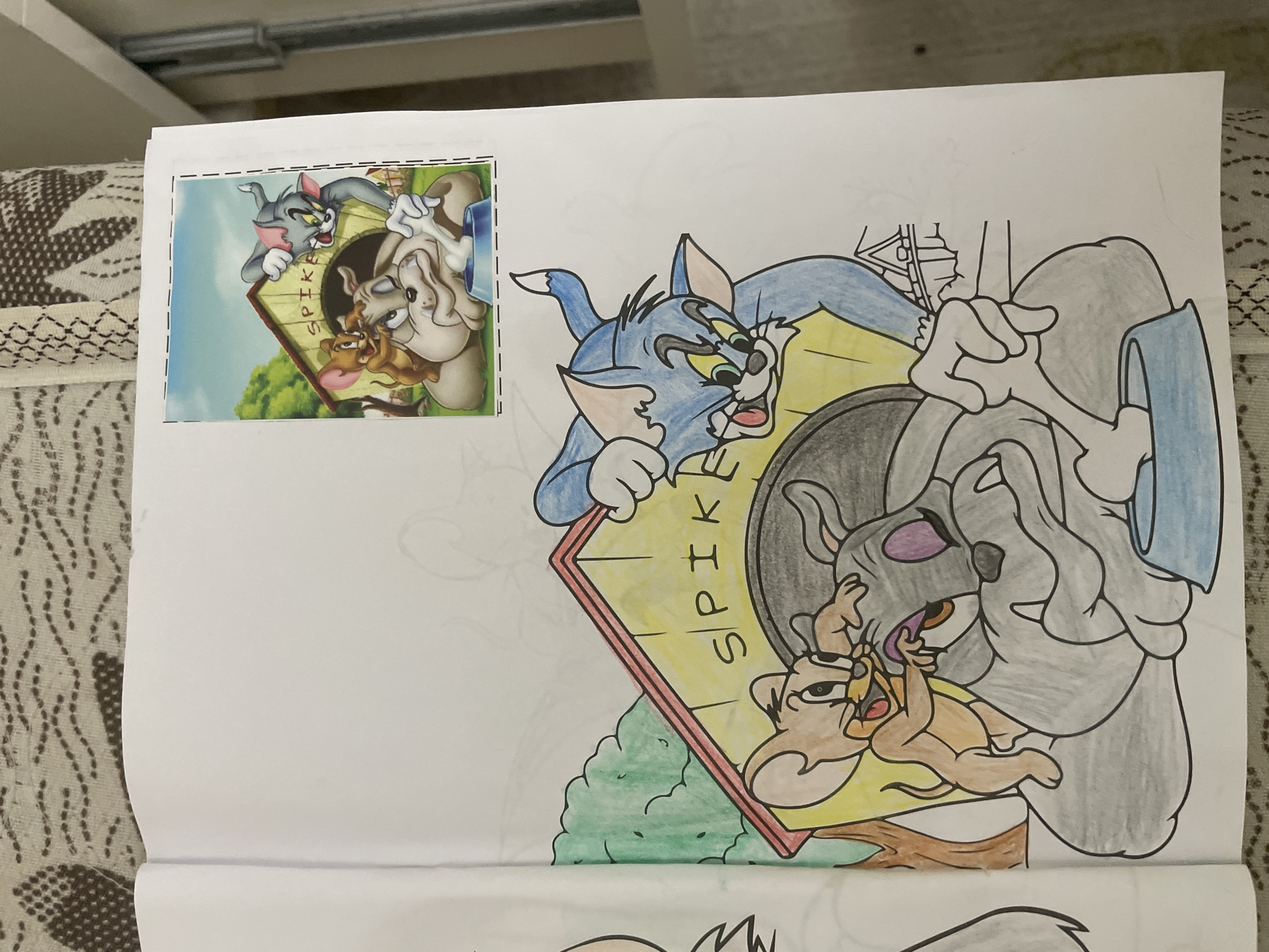 A physical artwork depicts a hand-colored drawing of characters from *Tom and Jerry*, accompanied by a smaller reference image. The main drawing is a partially colored rendering of Tom, Spike, and Jerry in a comical confrontation around Spike's doghouse.
Tom, the blue cat, is seen peeking over the top of a yellow doghouse, displaying an expression of distress or fear with wide eyes and an open mouth. Spike, the grey bulldog, emerges from the doghouse opening, looking surprised or annoyed. Jerry, the brown mouse, is clinging to Spike's head, appearing mischievous or triumphant. The word "SPIKE" is visibly written vertically on the side of the doghouse. A green bush is partially colored beneath the doghouse, suggesting an outdoor setting, while much of the background remains uncolored white space.
A smaller, full-color printed image of the identical cartoon scene is pasted onto the top-left corner of the page, serving as a reference. This smaller image shows the completed artwork with trees and a blue sky in the background.
The main drawing is colored with colored pencils; Tom is primarily blue and yellow, Jerry is brown with pink ears, and Spike is a light grey. The activity centers around the classic animated rivalry, with Tom seemingly caught in a predicament involving Spike and Jerry.
The photograph of this artwork was taken in Ihtiman, Bulgaria.