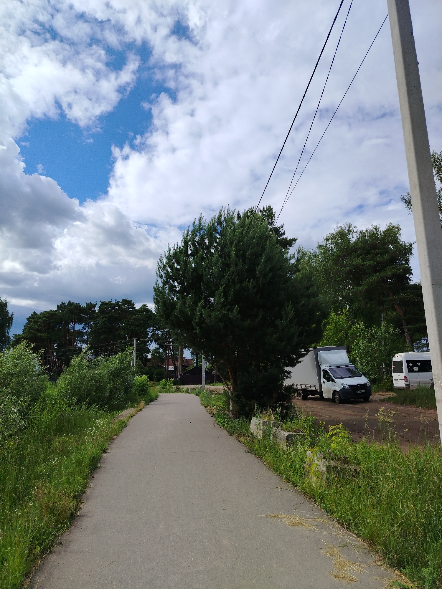 An asphalt pathway curves into the distance through a rural landscape under a partly cloudy sky. The pathway is flanked by tall, lush green grass and vegetation on the left, and a row of trees and shrubs on the right. In the middle ground, a white box truck with a tarped trailer is parked on a dirt area to the right of the pathway. Further to the right, a white minibus is also parked. A large evergreen tree stands prominently on the right side of the pathway in the foreground. In the background, a line of pine trees is visible, with some residential structures interspersed. A concrete utility pole stands to the far right of the image. The overall scene suggests a peaceful, possibly semi-urban or suburban environment in the city of Kimry, Russia. There are no people visible in the image.