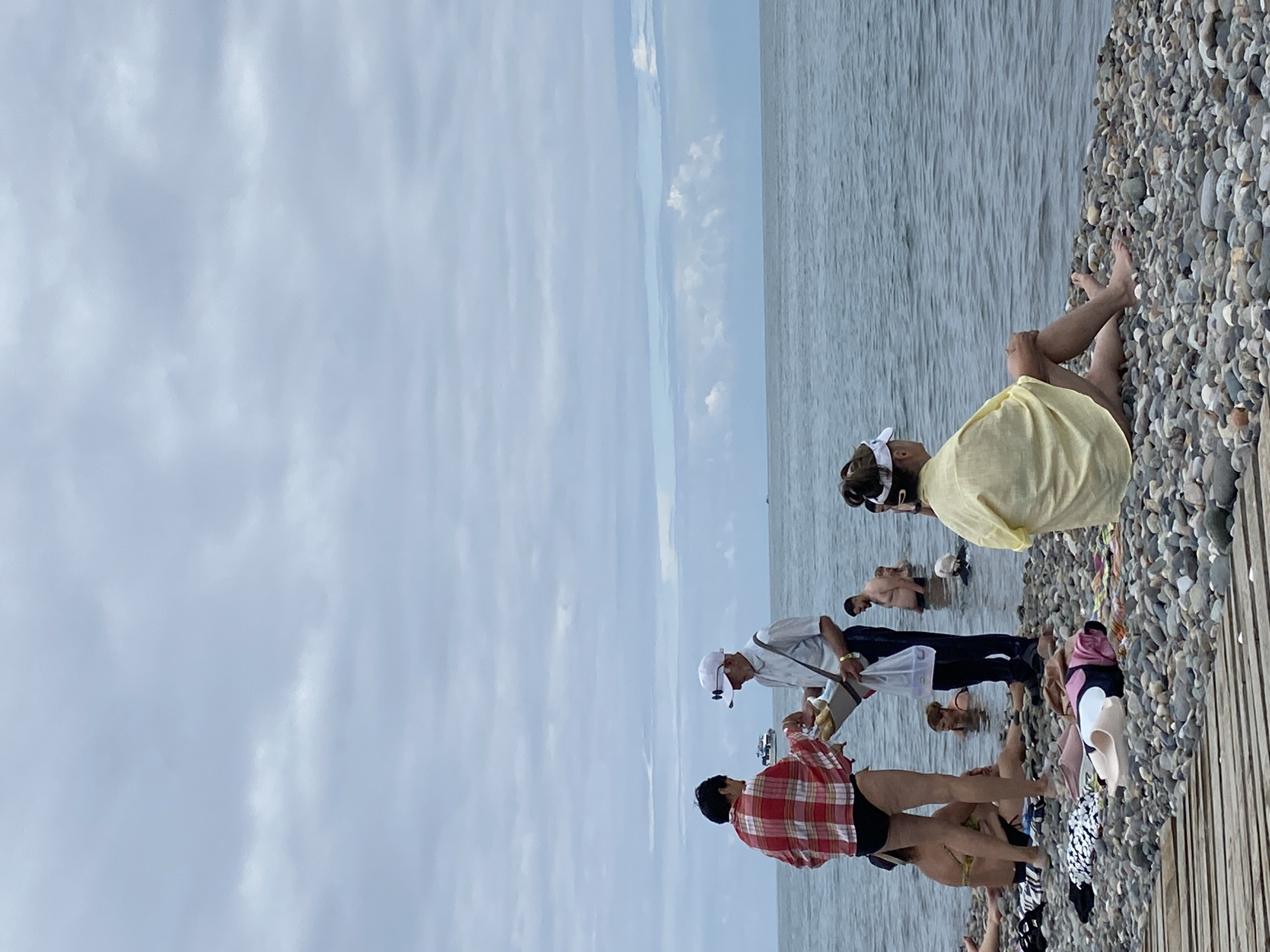 The image depicts a cloudy day at a pebble beach in Batumi, Georgia. In the foreground, a woman in a yellow shawl sits on the pebbles, facing the sea. To her left, a wooden boardwalk is partially visible. A man in a white shirt and cap stands in the shallow water, holding a plastic bag filled with items and interacting with a woman in a red and white checkered shirt. Several other people are in the water, some swimming and others standing. The sea is calm, and a boat is visible in the distance. The overall atmosphere is relaxed and casual, typical of a beach day.