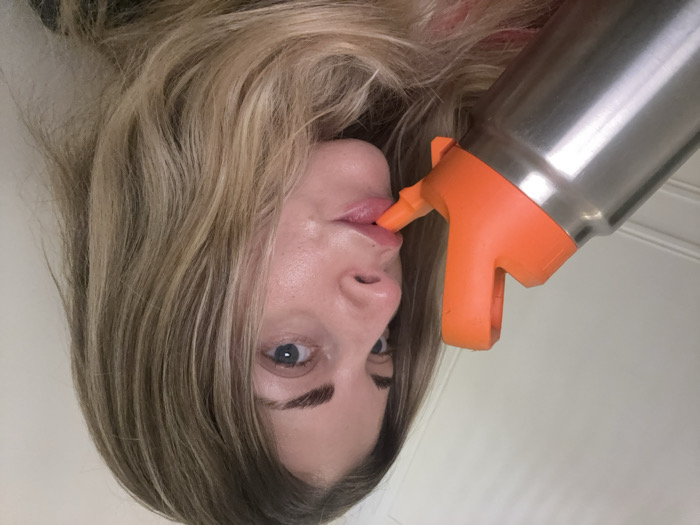 A female individual with light blonde and brown wavy hair and blue eyes is captured in a close-up, slightly angled shot. She is actively drinking from a stainless steel insulated water bottle, whose bright orange flip-top lid and straw spout are positioned against her lips. Her gaze is directed towards the viewer. The immediate environment is an indoor setting with a plain, light-colored wall forming the background. A subtle white decorative molding or trim is visible on the wall to the left. The scene occurs indoors, within a room located in Paris, France.