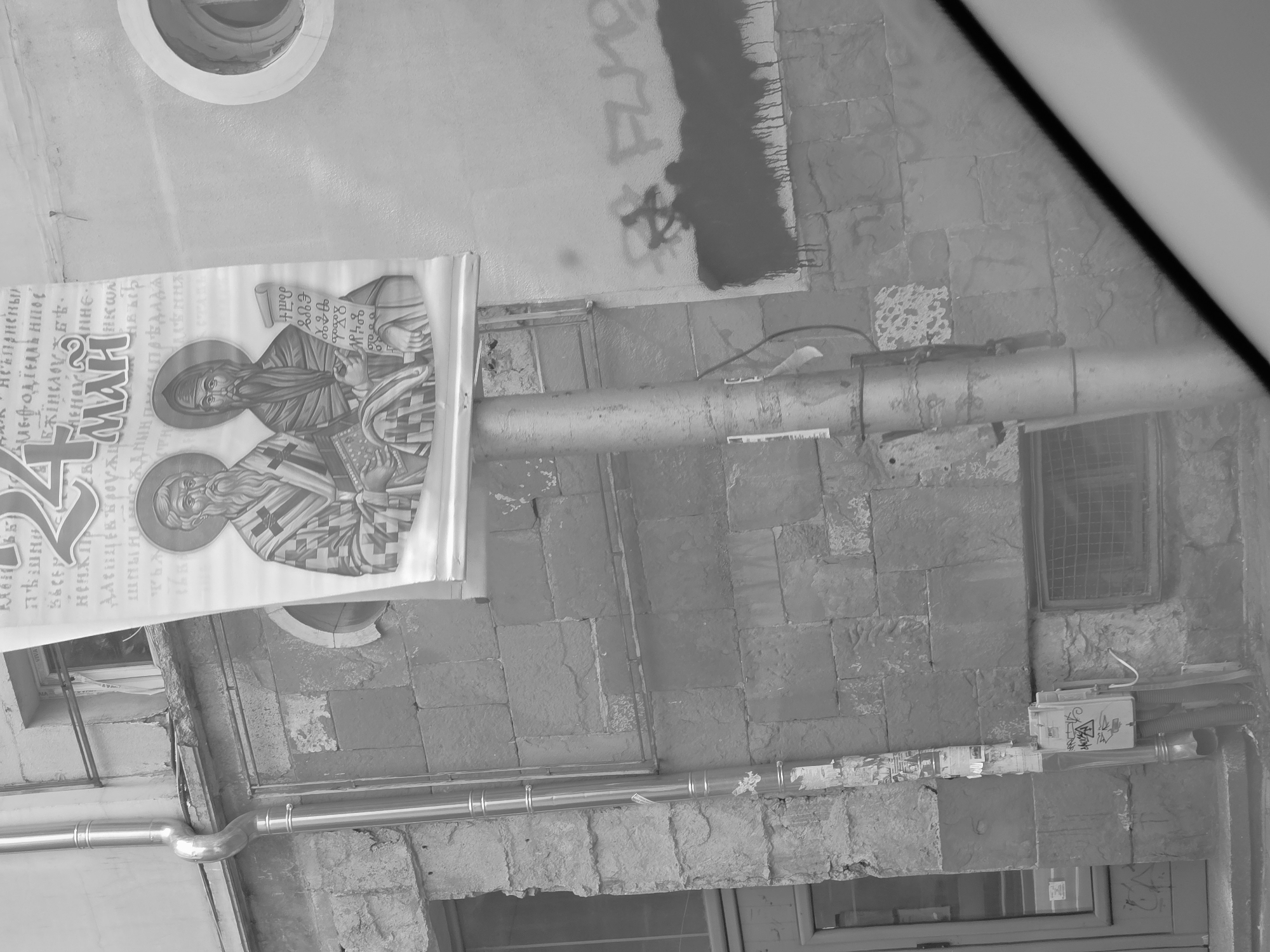 The image shows an exterior wall with various features. A poster depicting two religious figures is affixed to the upper left. A weathered, gray pipe runs horizontally across the middle of the wall. A barred window is visible on the right. Graffiti marks a section of the wall above the pipe. A utility box with further graffiti is located in the lower right. The wall is constructed of stone blocks. The image is likely taken in Varna, Bulgaria due to the religious imagery and architectural style.