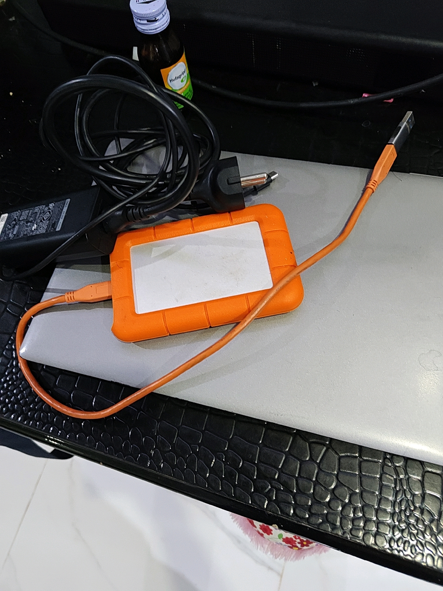 An overhead shot shows a black power cord coiled on a black surface. Next to the cord is an orange external hard drive connected to an orange USB cable. The cable runs to the right, and its other end connects to a black laptop. A power adapter with a black cord is partially visible behind the coiled cord. A small bottle with a white cap and a green and yellow label is behind the coiled cord. The scene appears to be indoors, on a black textured surface.