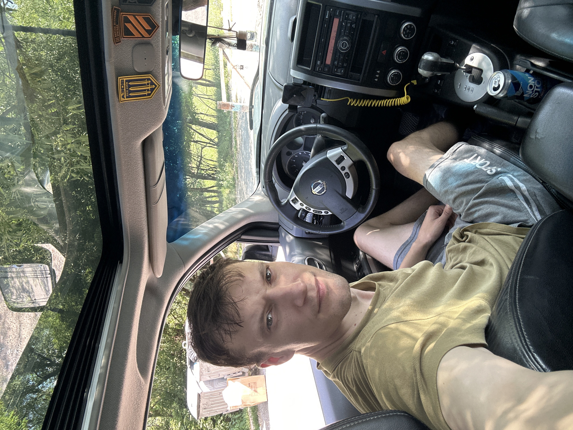 A man sits in the driver's seat of a car, with a panoramic sunroof open above him. He is wearing a tan t-shirt and grey athletic shorts. The car's dashboard is visible, featuring a Nissan steering wheel, a radio, and a gear shifter. A smartphone is mounted on the dashboard. Two small patches are affixed to the car's interior overhead console. Outside the car, two large gray trash bins and a cardboard box are visible next to a tree-lined road. The scene appears to be an outdoor location, possibly in Makiv, Ukraine, given the presence of Ukrainian military-style patches.