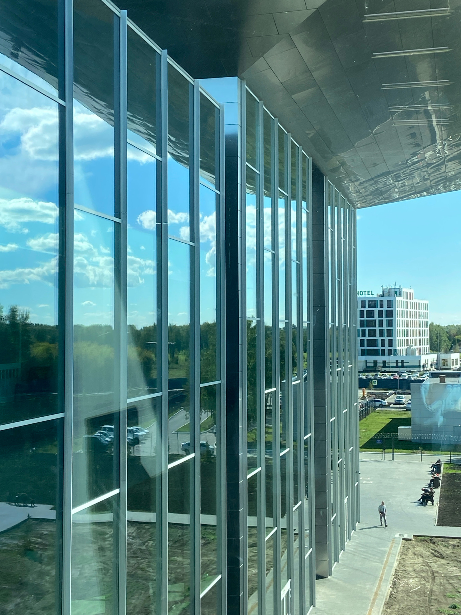 The image captures an exterior view of a modern building with a glass facade in the city of Ob, Russia. The building's reflective glass windows show a clear blue sky with white clouds and the surrounding landscape, which includes trees and other buildings. In the distance, a multi-story building with "HOTEL" signage is visible, along with a parking lot filled with cars. The foreground features a paved walkway with a few people seated on benches and one person walking. A section of the building's dark, textured roof overhangs the glass facade. The overall scene depicts a sunny day in an urban or suburban setting.