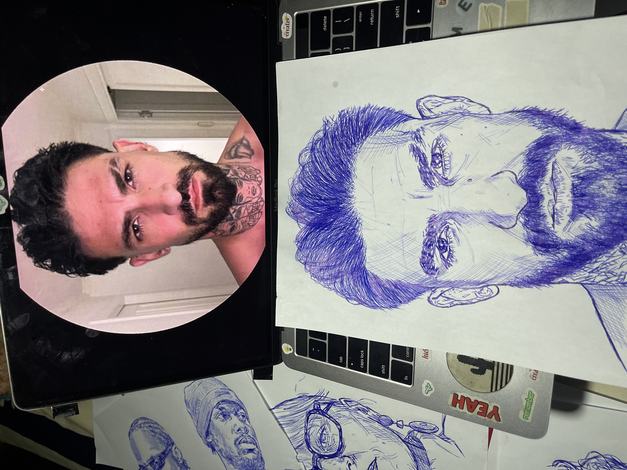 The image depicts a close-up of a laptop screen displaying a portrait of a man with tattoos. In front of the laptop, a hand-drawn sketch of the same man, also with tattoos, is laid out. Several other sketches of faces are visible on the table, some drawn with blue ink. The laptop is open, and the keyboard is visible. The overall scene suggests an artist at work, using a digital image as reference for their drawing. There is no indication of the location being Jos, Nigeria, as the image does not contain any recognizable landmarks or contextual clues to suggest this specific city.