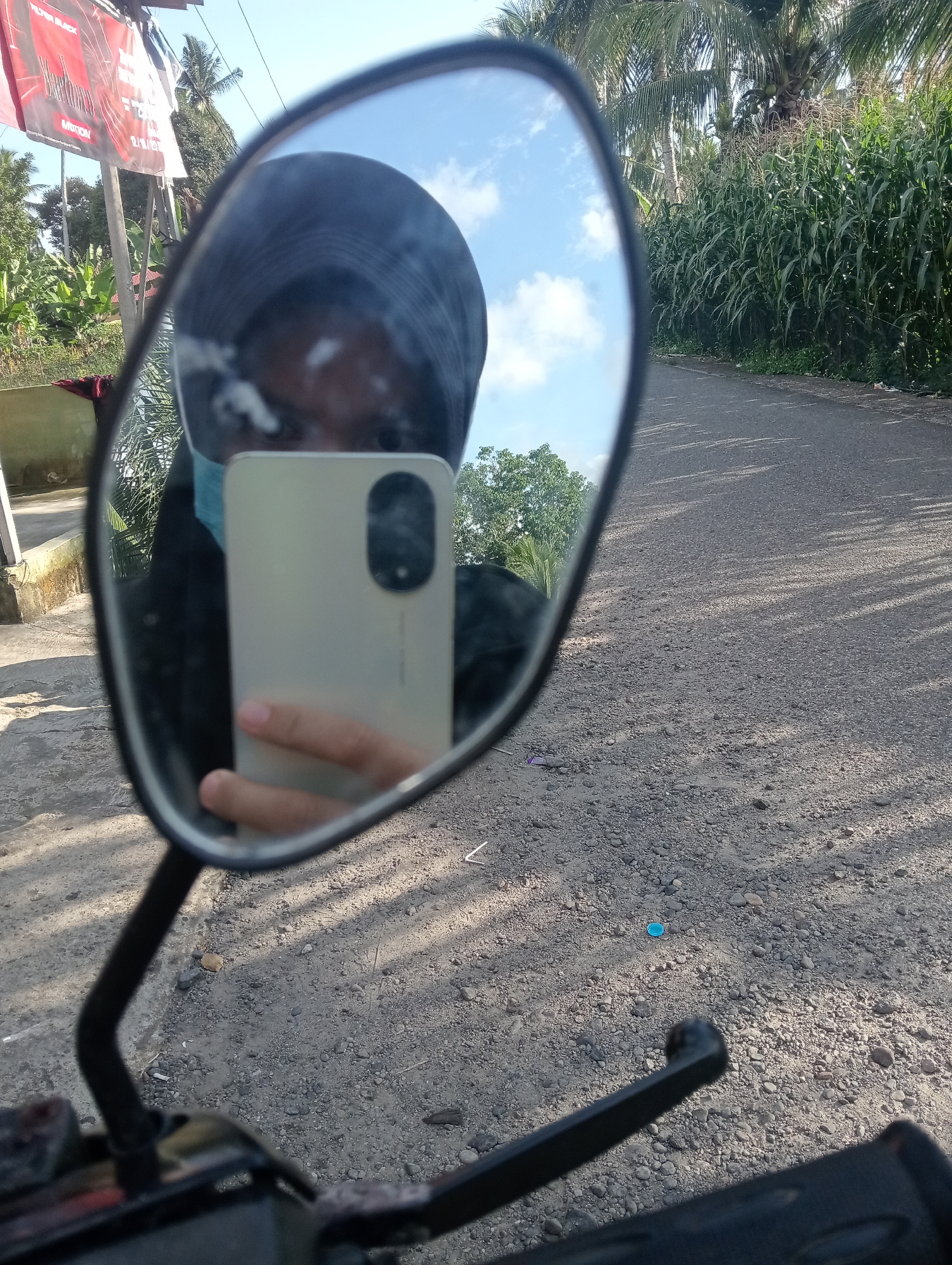 A person wearing a headscarf and a light blue mask is reflected in a motorcycle's rearview mirror. They are holding a light-colored smartphone in their right hand, taking a selfie. The reflection also shows the upper part of the person's body, including a dark jacket. The background in the mirror shows a road with gravel, green corn stalks lining the right side, and palm trees and a blue sky with clouds. The location is identified as Campago, Indonesia. The foreground of the image shows the motorcycle's handlebars and part of the mirror assembly.
